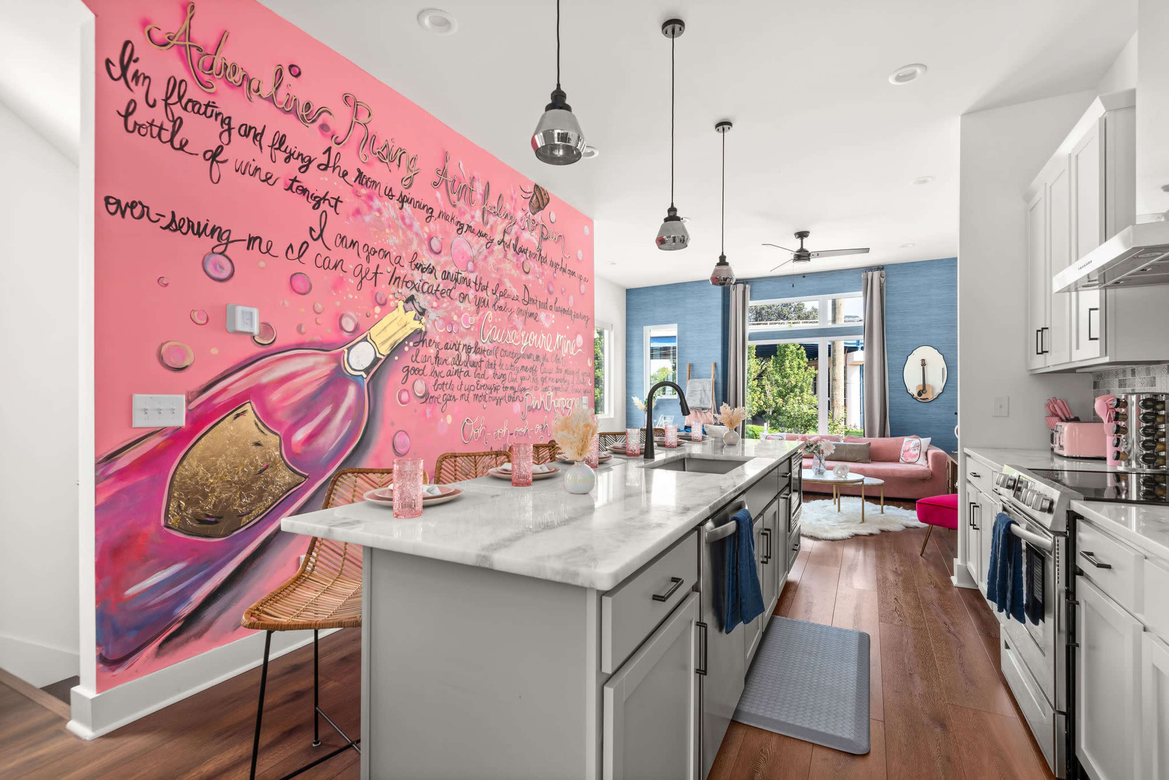 A modern kitchen features a pink mural of a wine bottle, with a marble countertop and a view into a brightly furnished living area.