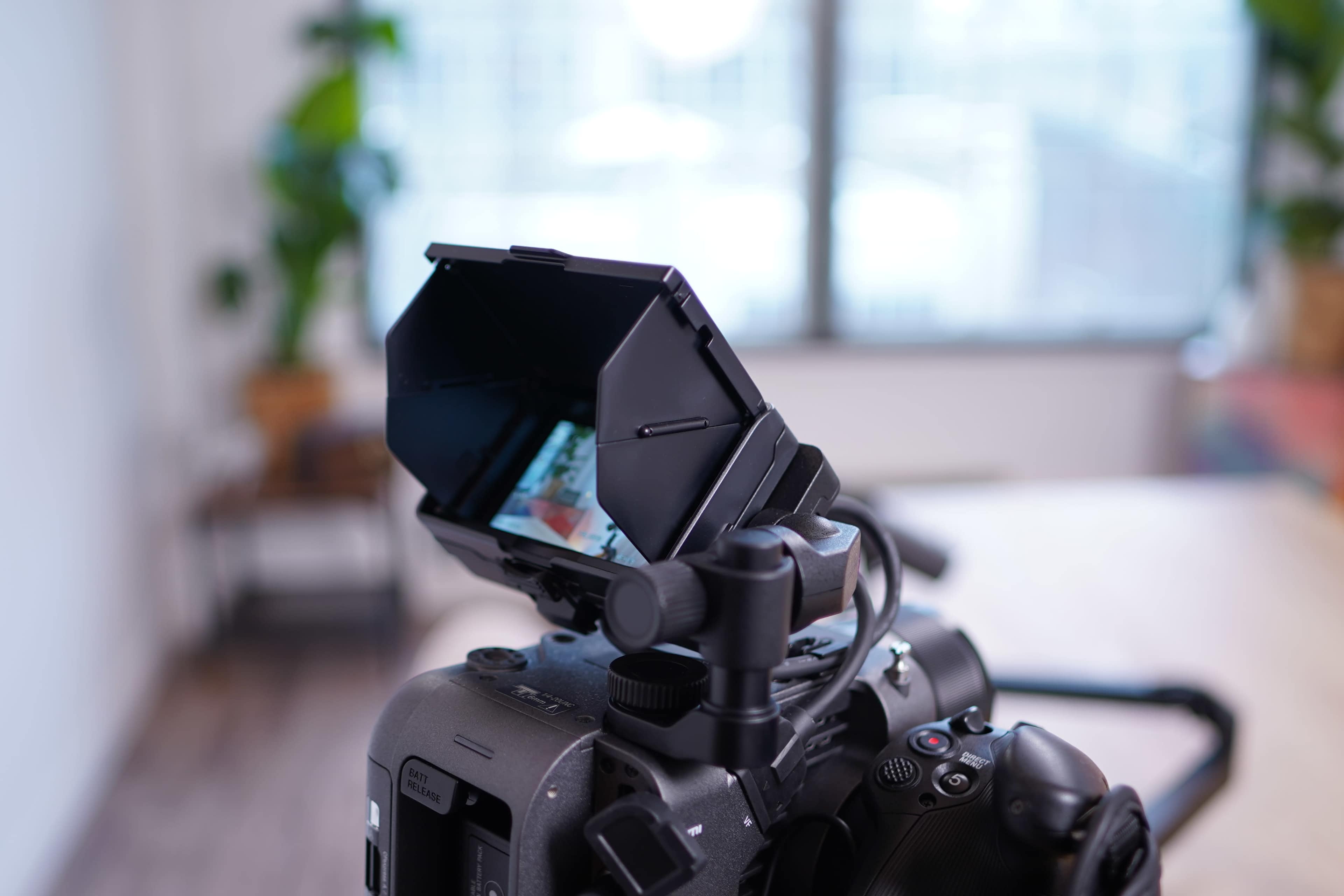 A camera with a monitor hood is positioned on a tripod, focusing on a blurred background featuring greenery.