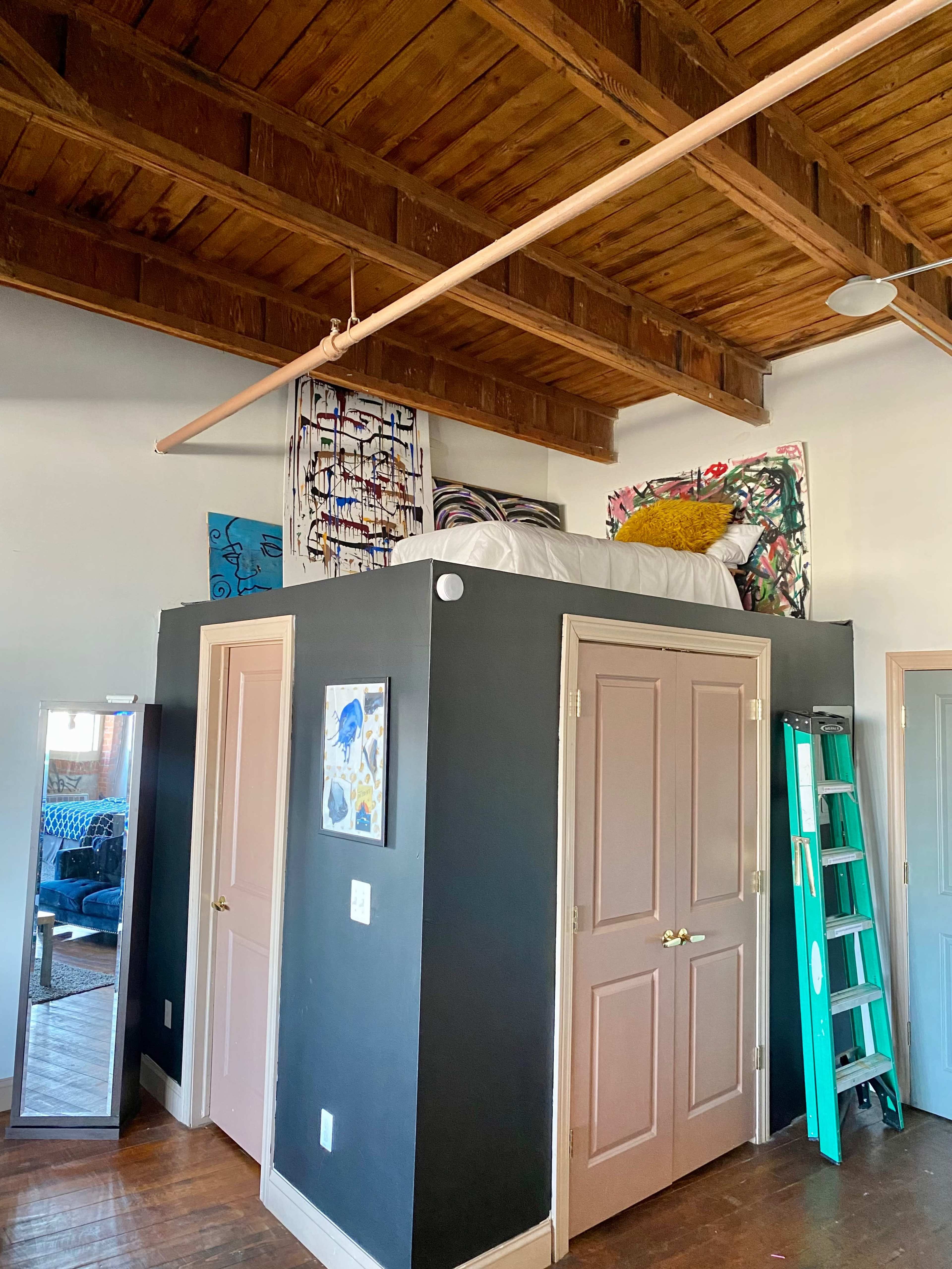 A small room with a raised bed area above two closets, featuring wooden beams overhead and colorful artwork on the walls.