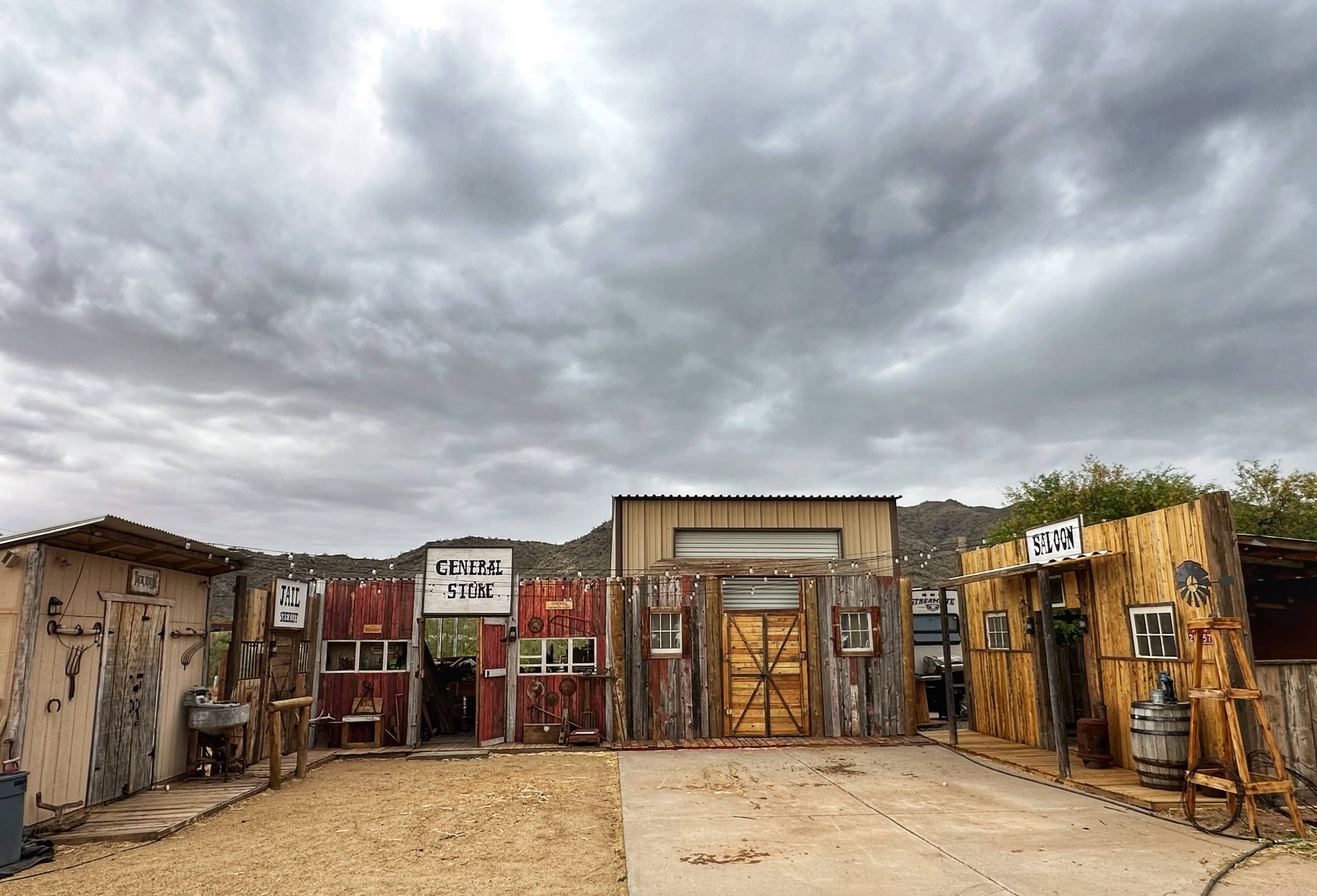 A rustic facade featuring several weathered wooden structures labeled "General Store" and "Saloon" under a cloudy sky.