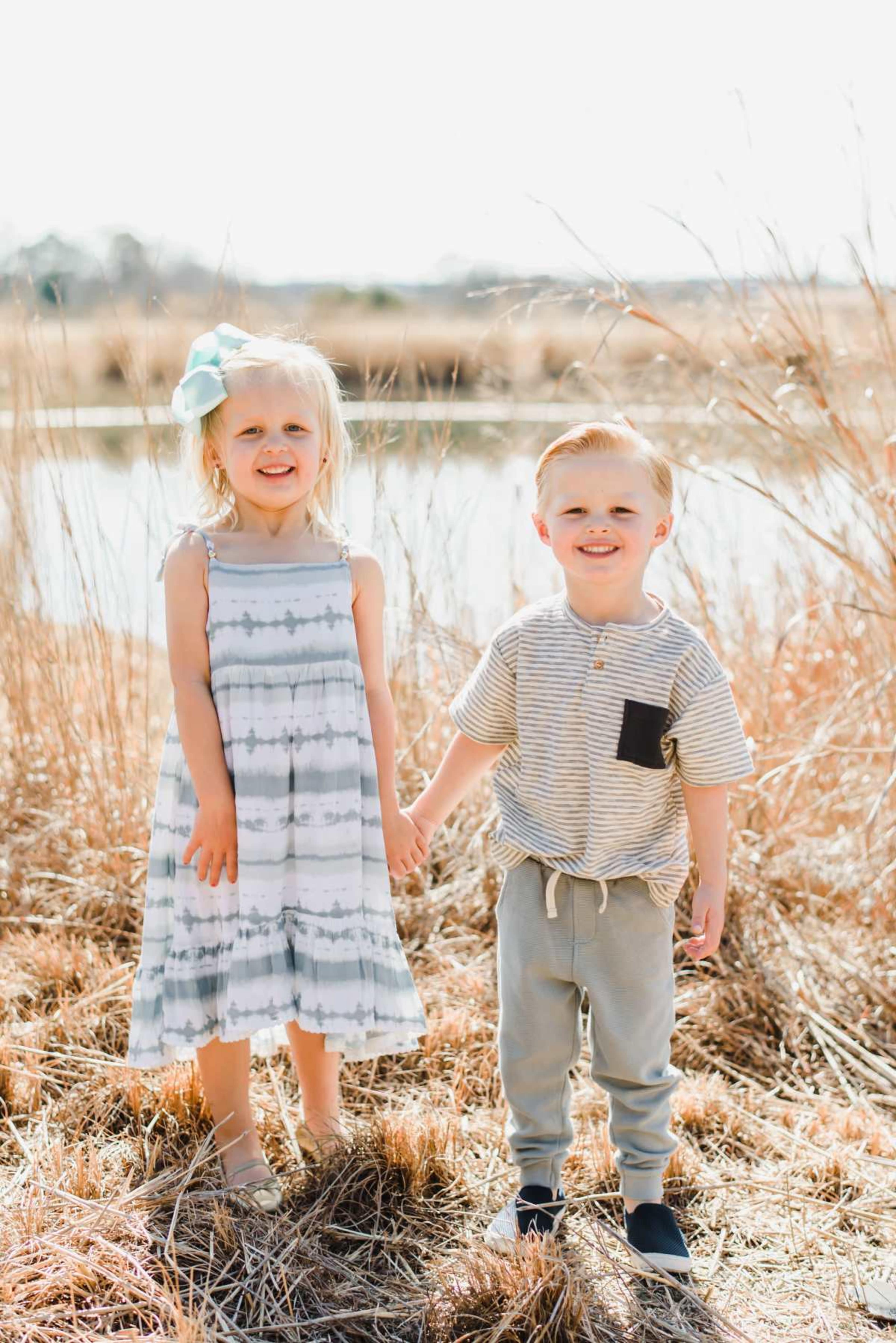 Two young children stand hand in hand in a grassy field near a body of water, with dry plants surrounding them.