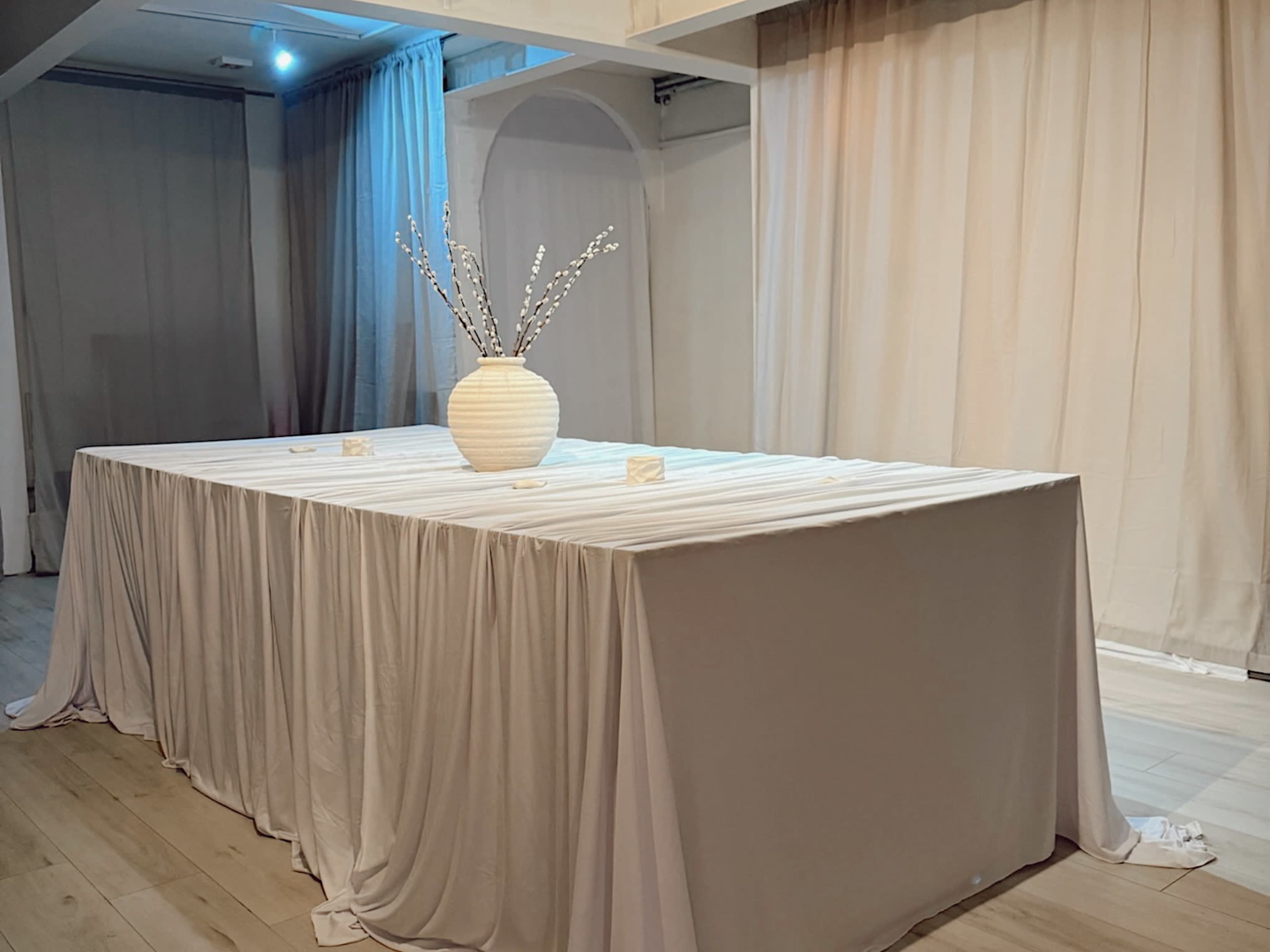 A large, elegantly draped table is set in a softly lit room, featuring a simple vase with decorative branches at its center.