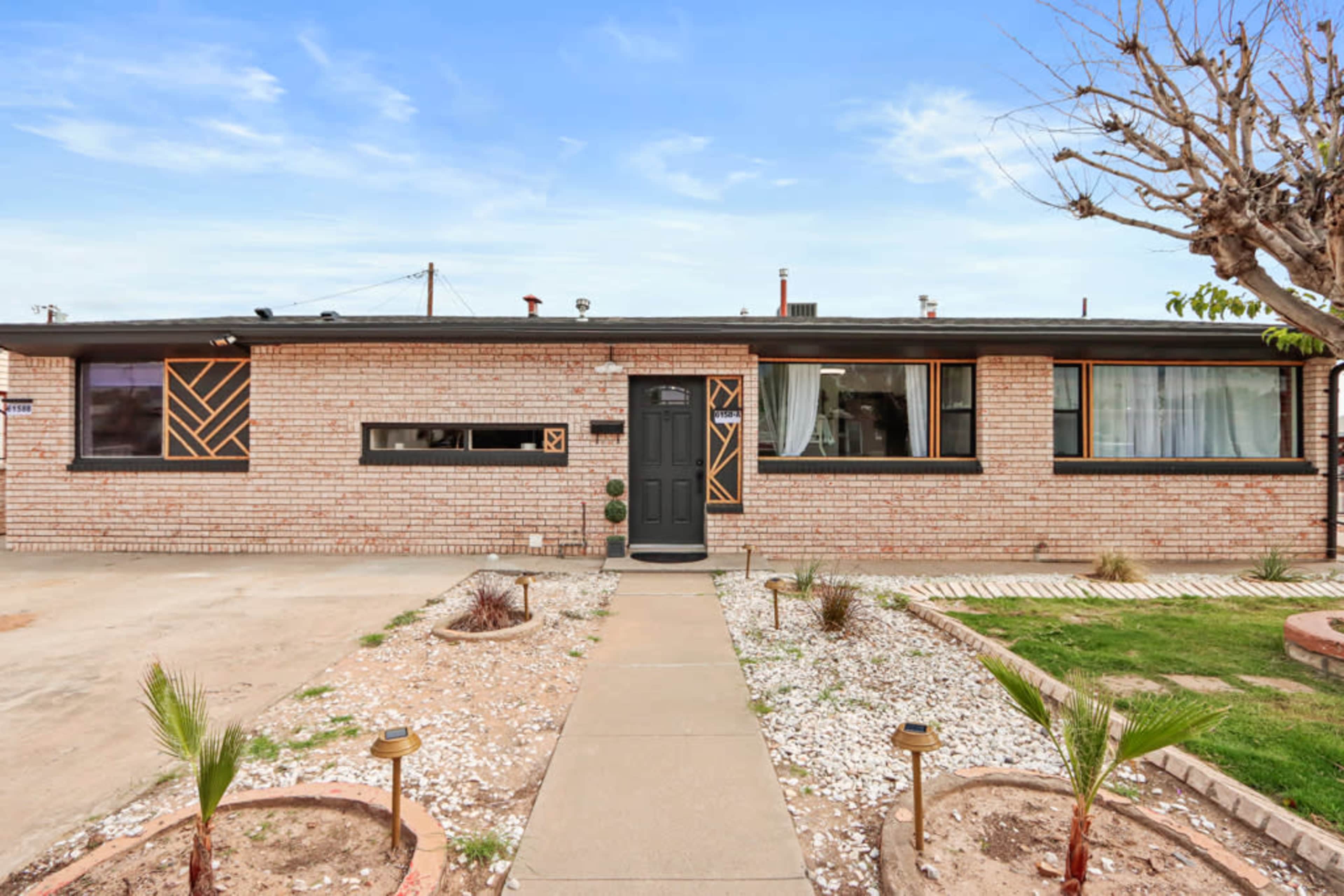 A single-story brick house with a front pathway, surrounded by gravel and sparse landscaping, under a clear blue sky.