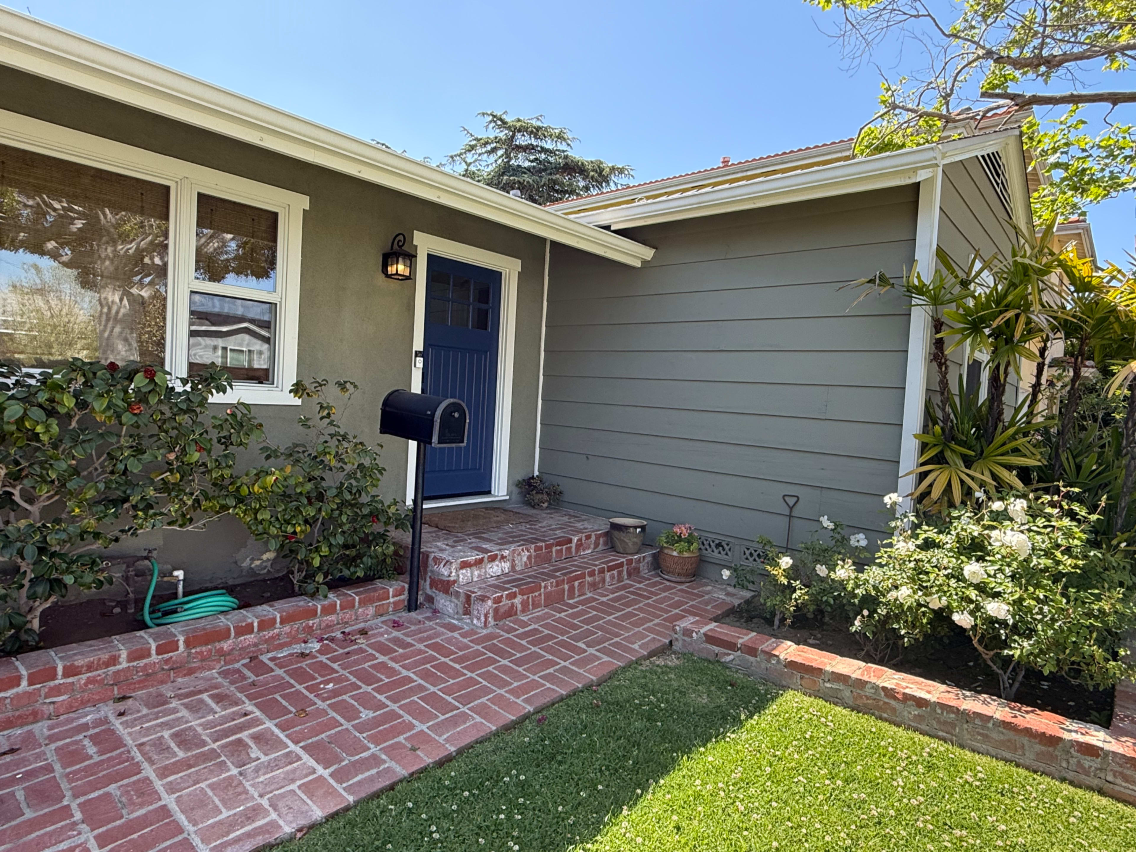 The image shows a front yard of a house with a blue door, brick steps, and landscaped greenery.
