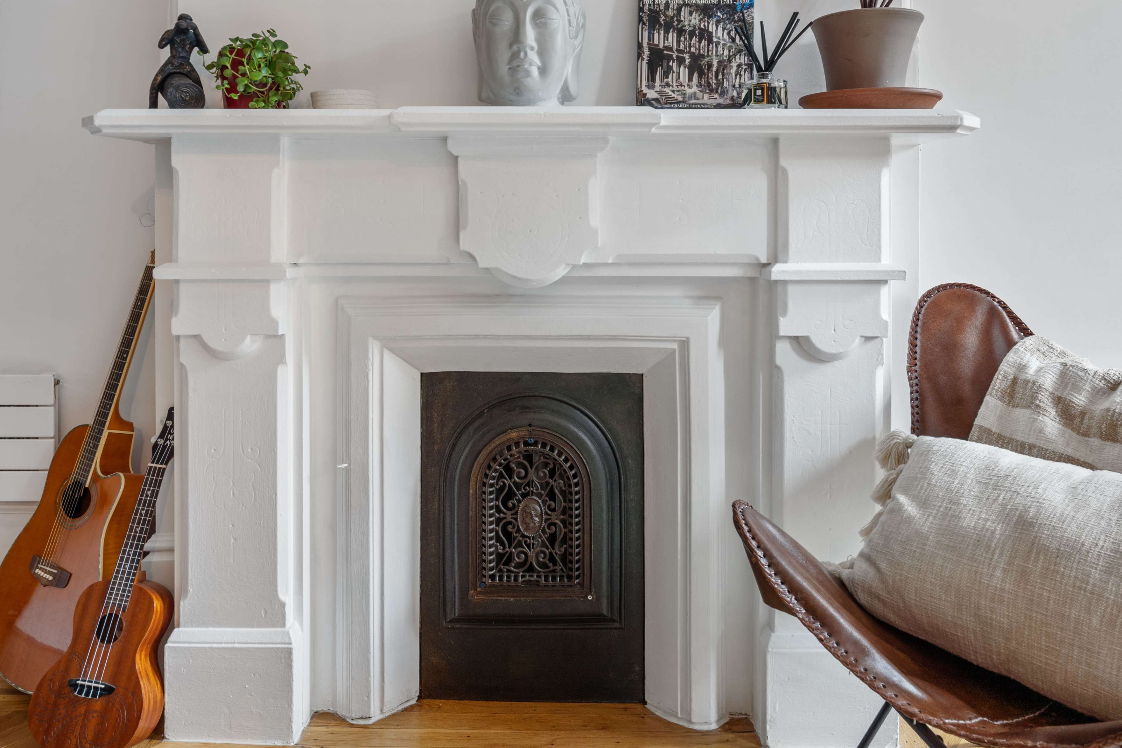 The image features a white fireplace adorned with a decorative statue, surrounded by a guitar, potted plants, and a chair with cushions.