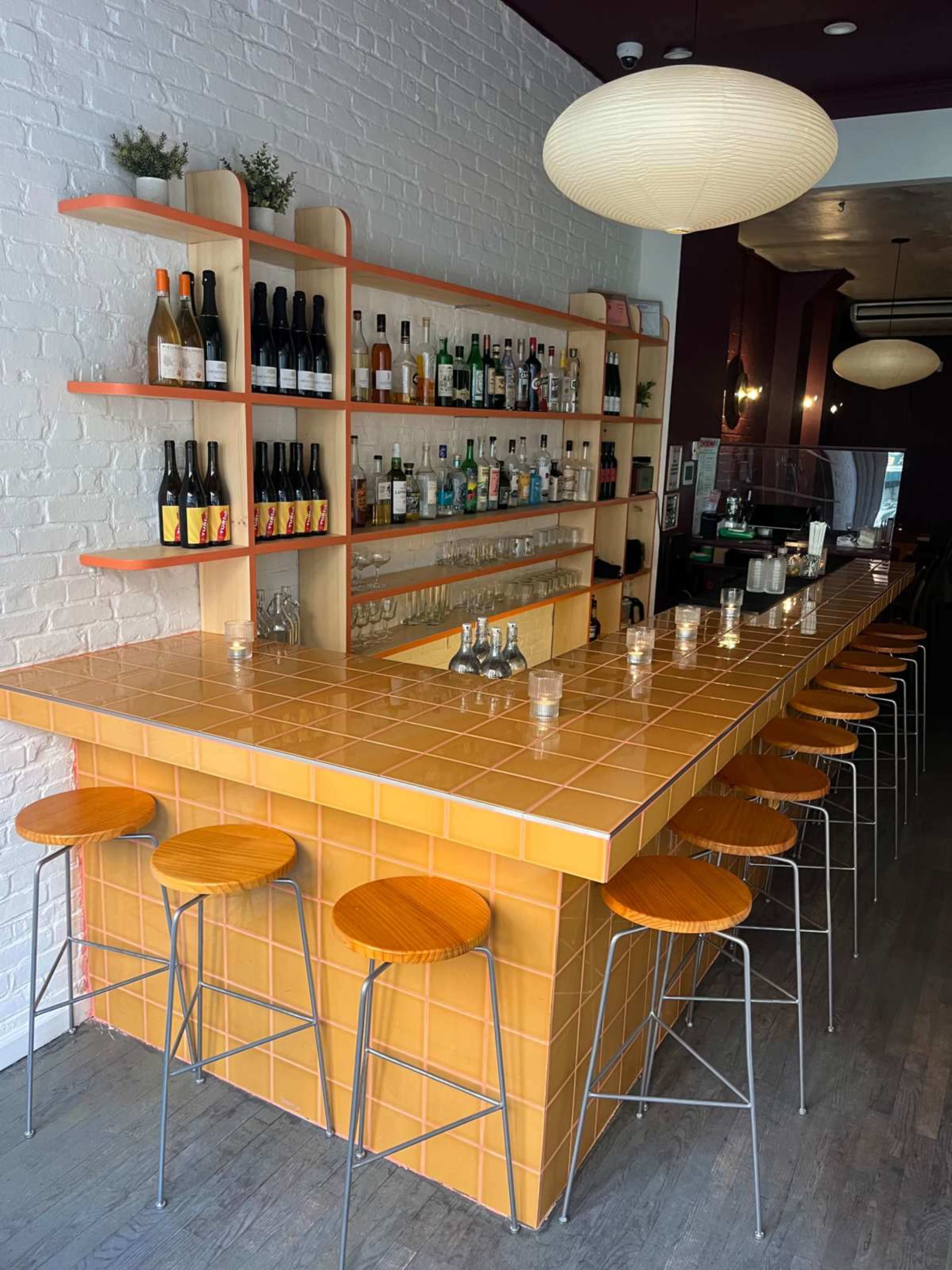 The image shows a brightly colored bar with an orange and yellow tiled countertop and a wooden back shelf displaying various bottles.