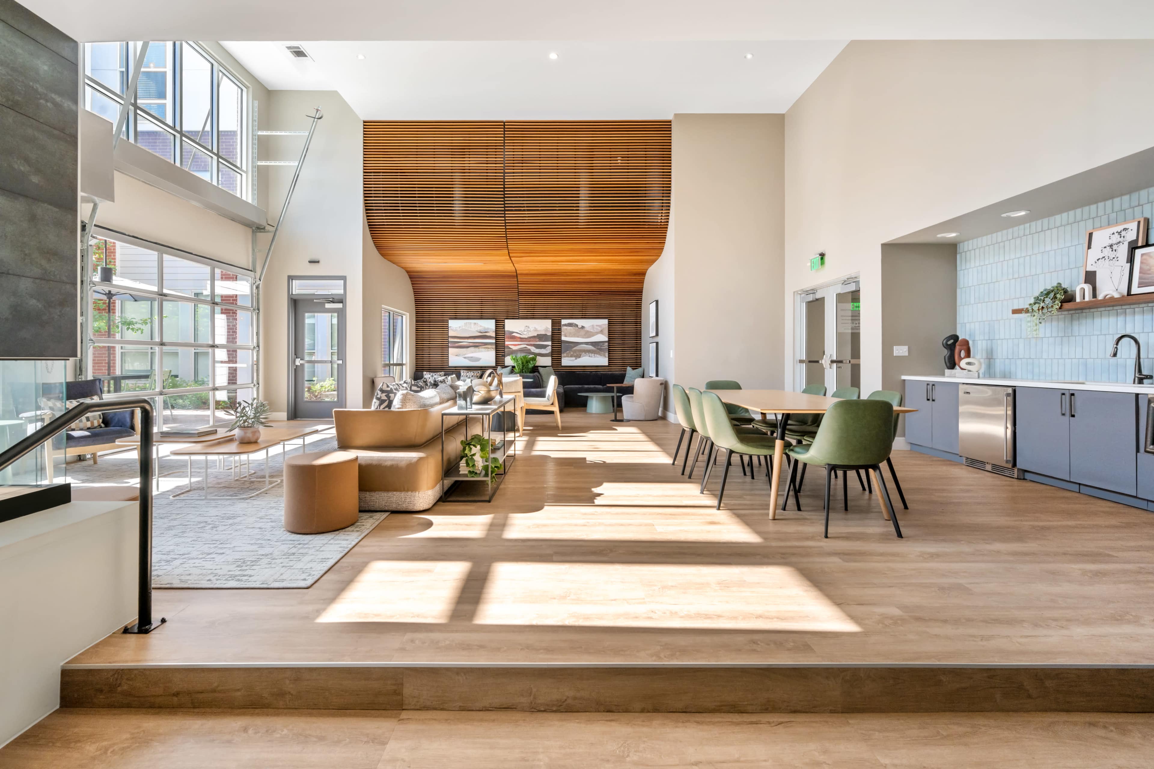 The image shows a spacious, modern communal area featuring seating arrangements, large windows allowing natural light, and a kitchen area in the background.