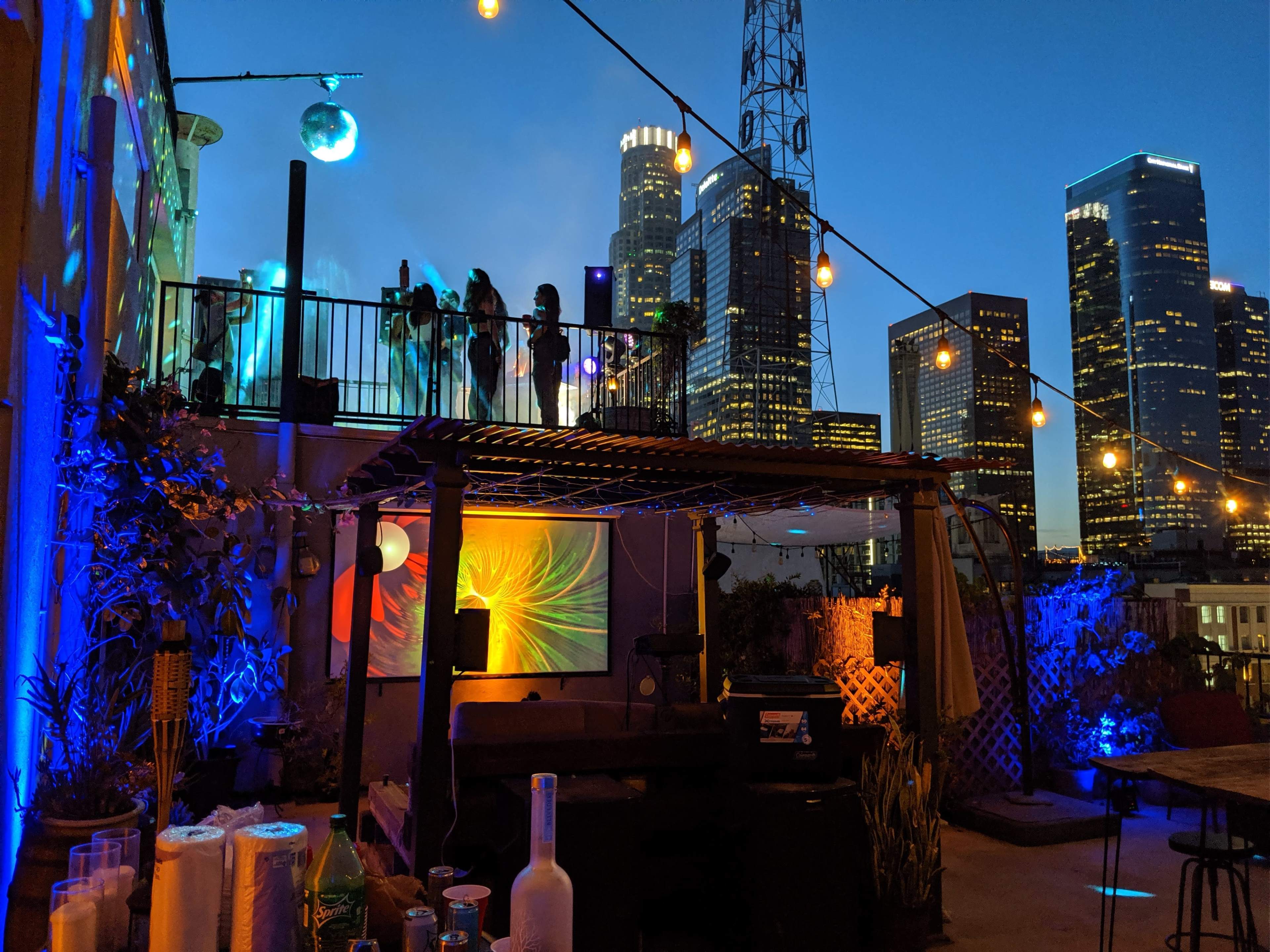 A rooftop bar with colorful lighting and city skyscrapers in the background at dusk.