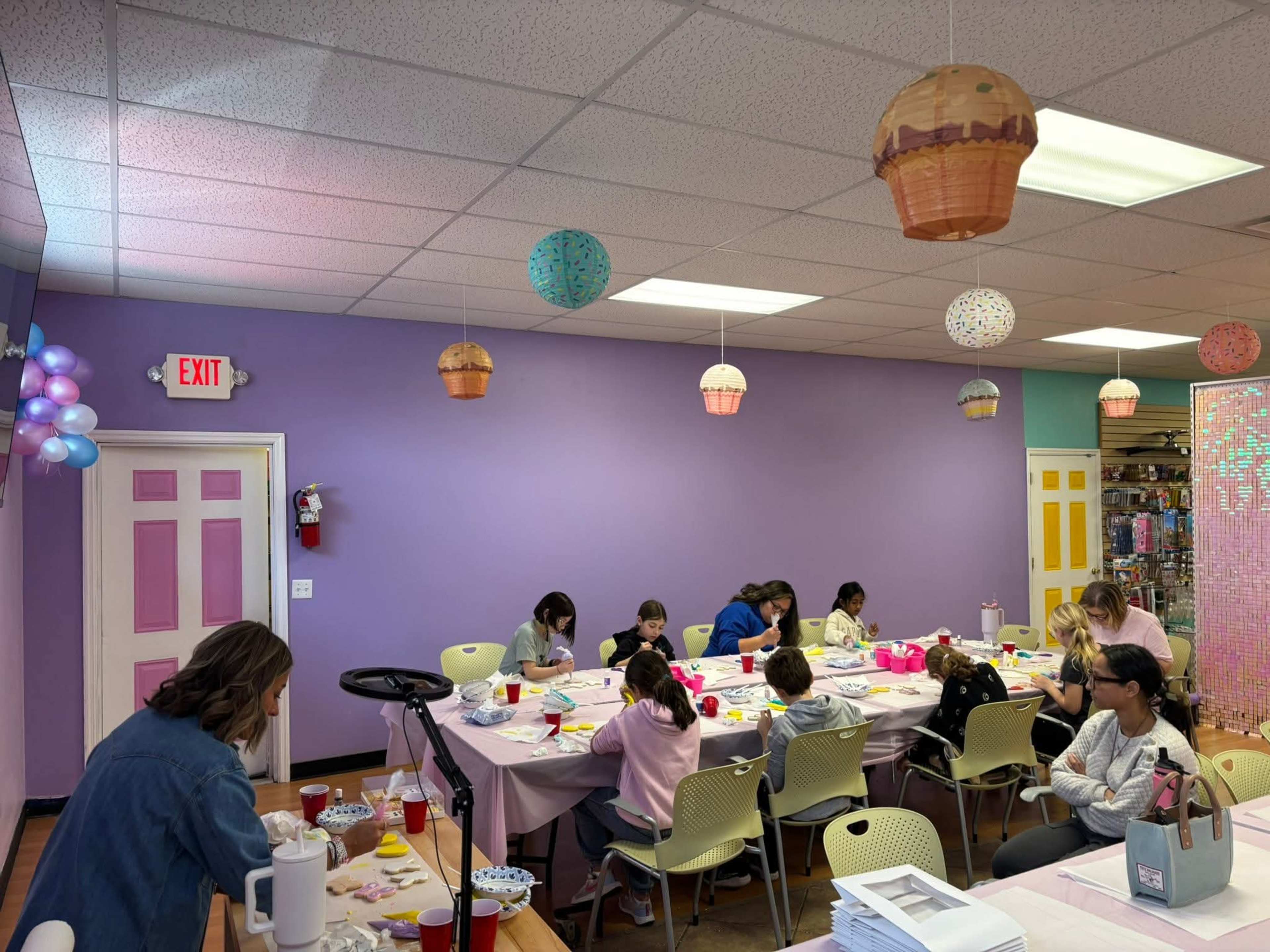 The image shows a colorful craft room where several children and adults participate in a creative activity, with tables set up for decorating cookies and hanging decorative lanterns above.