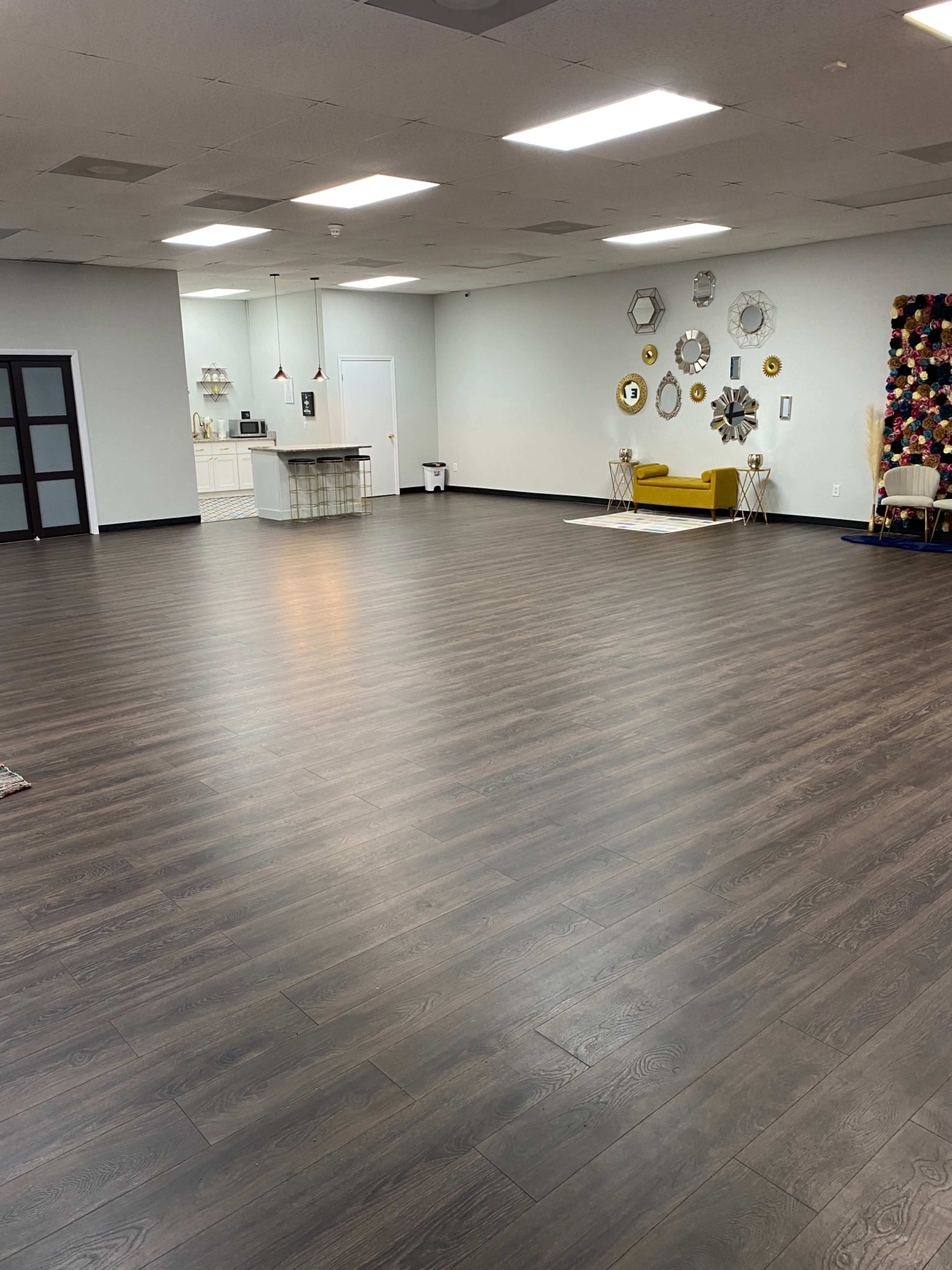 The image shows a spacious, empty room with dark wooden flooring, a kitchenette in the background, and decorative wall hangings on one side.