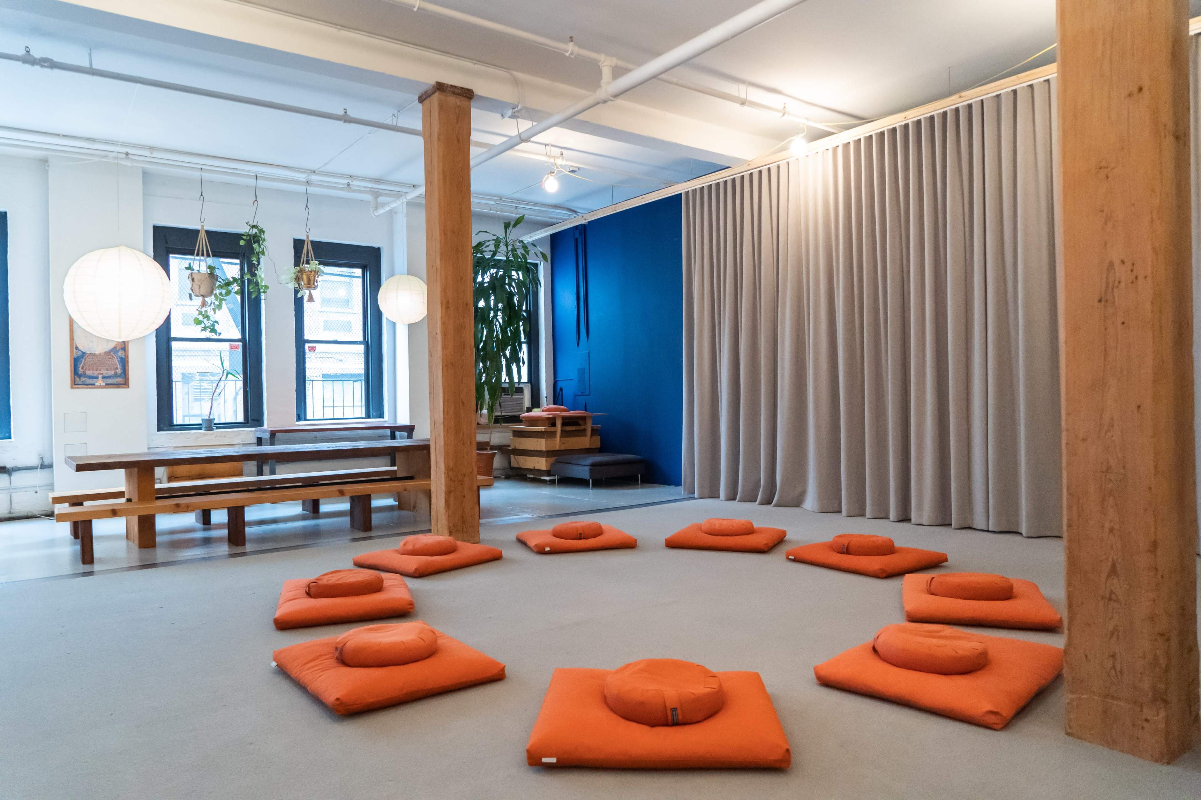 A meditation space with orange cushions arranged in a circular formation on a light-colored floor, surrounded by wooden beams and a curtain.
