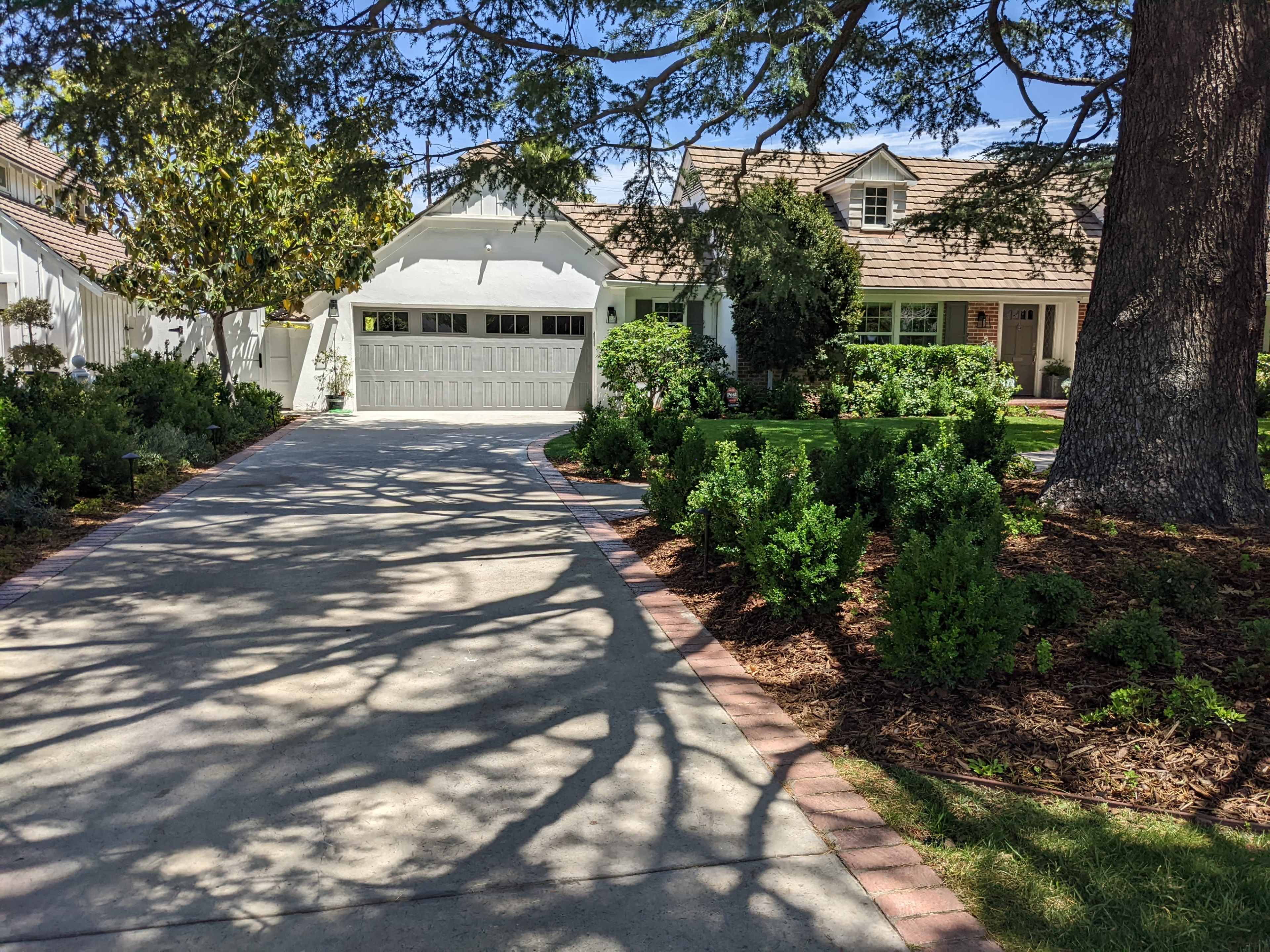 A driveway leads to a house with a garage, surrounded by neatly trimmed bushes and trees casting shadows on the pavement.