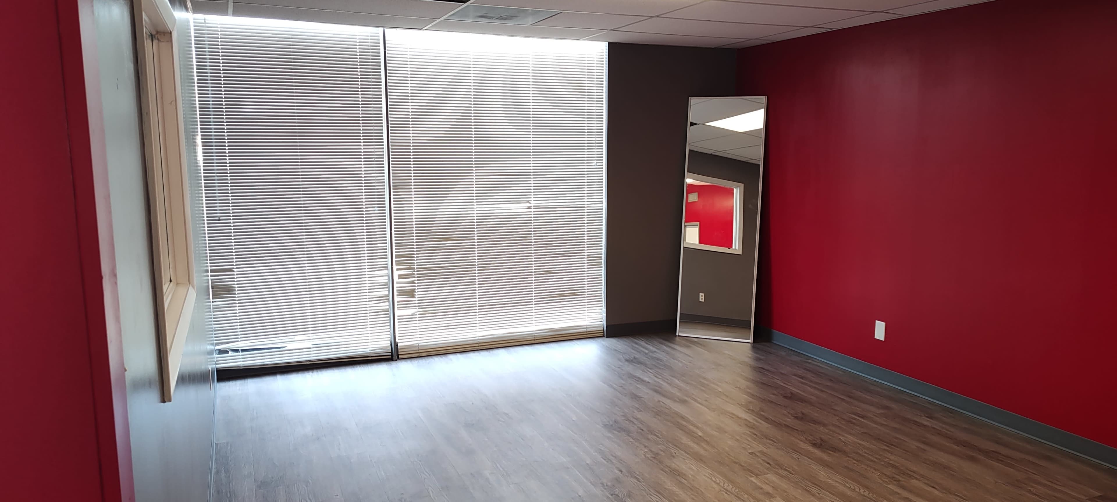 The image shows a room with red walls, a large window covered by blinds, a full-length mirror on one wall, and a wooden floor.