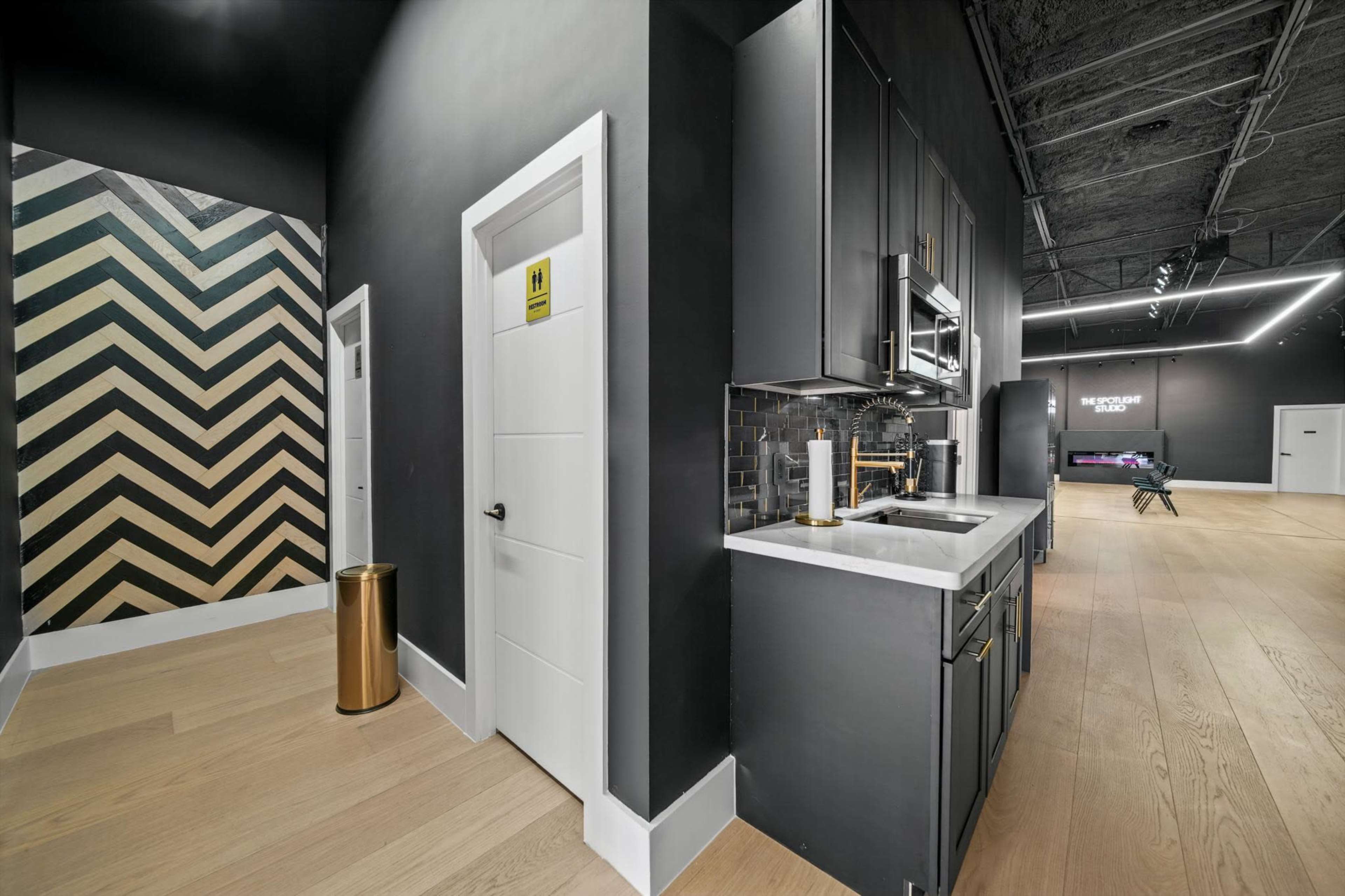 The image shows a modern interior space featuring a small kitchen area with dark cabinetry and a patterned accent wall, along with light wooden flooring.
