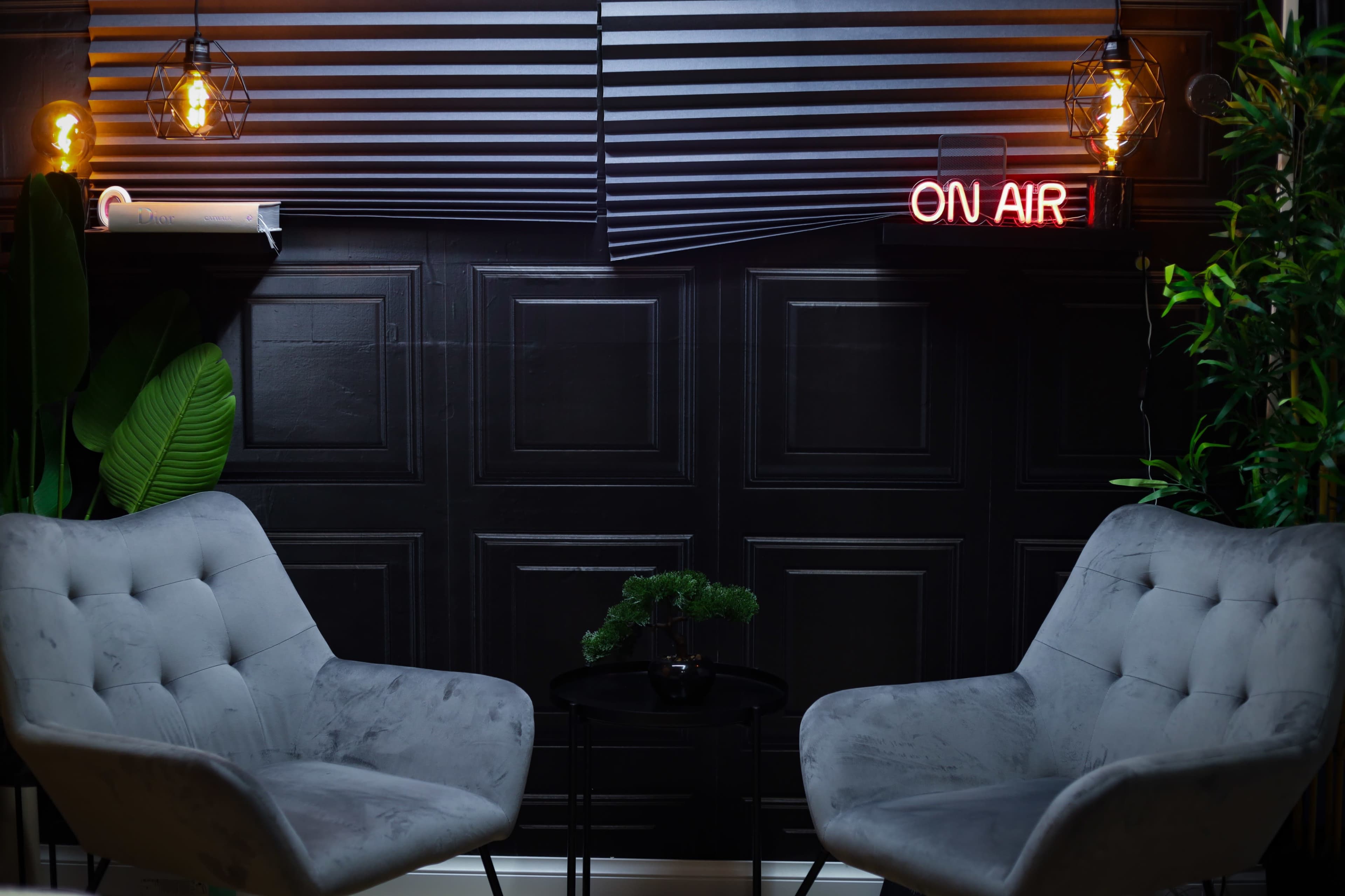 The scene features two gray armchairs facing a small black table, with a neon "ON AIR" sign and a pair of pendant lights illuminating a textured black wall.