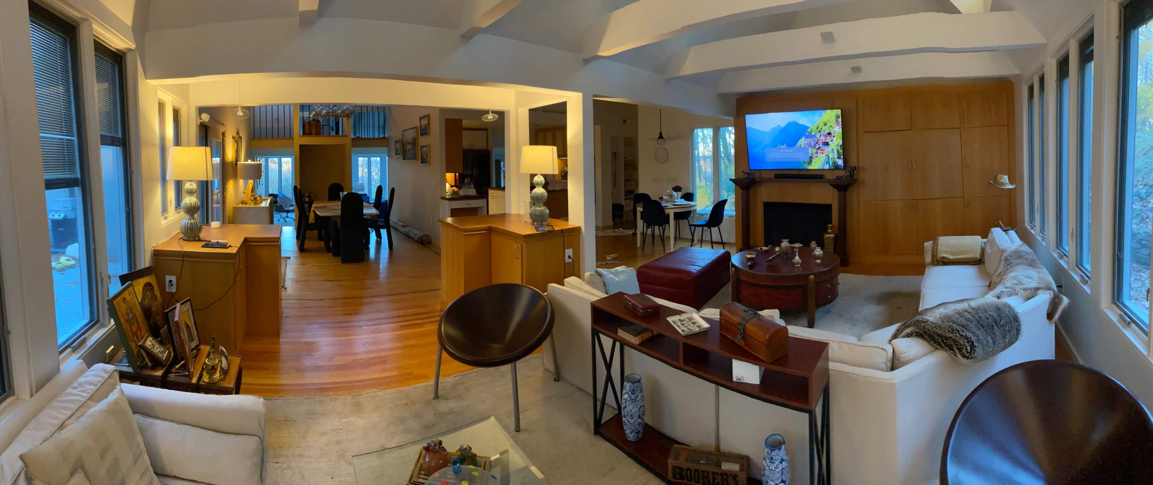 A spacious living room features a white sectional sofa, a wooden coffee table, and a large television displaying a landscape, with a dining area visible in the background.
