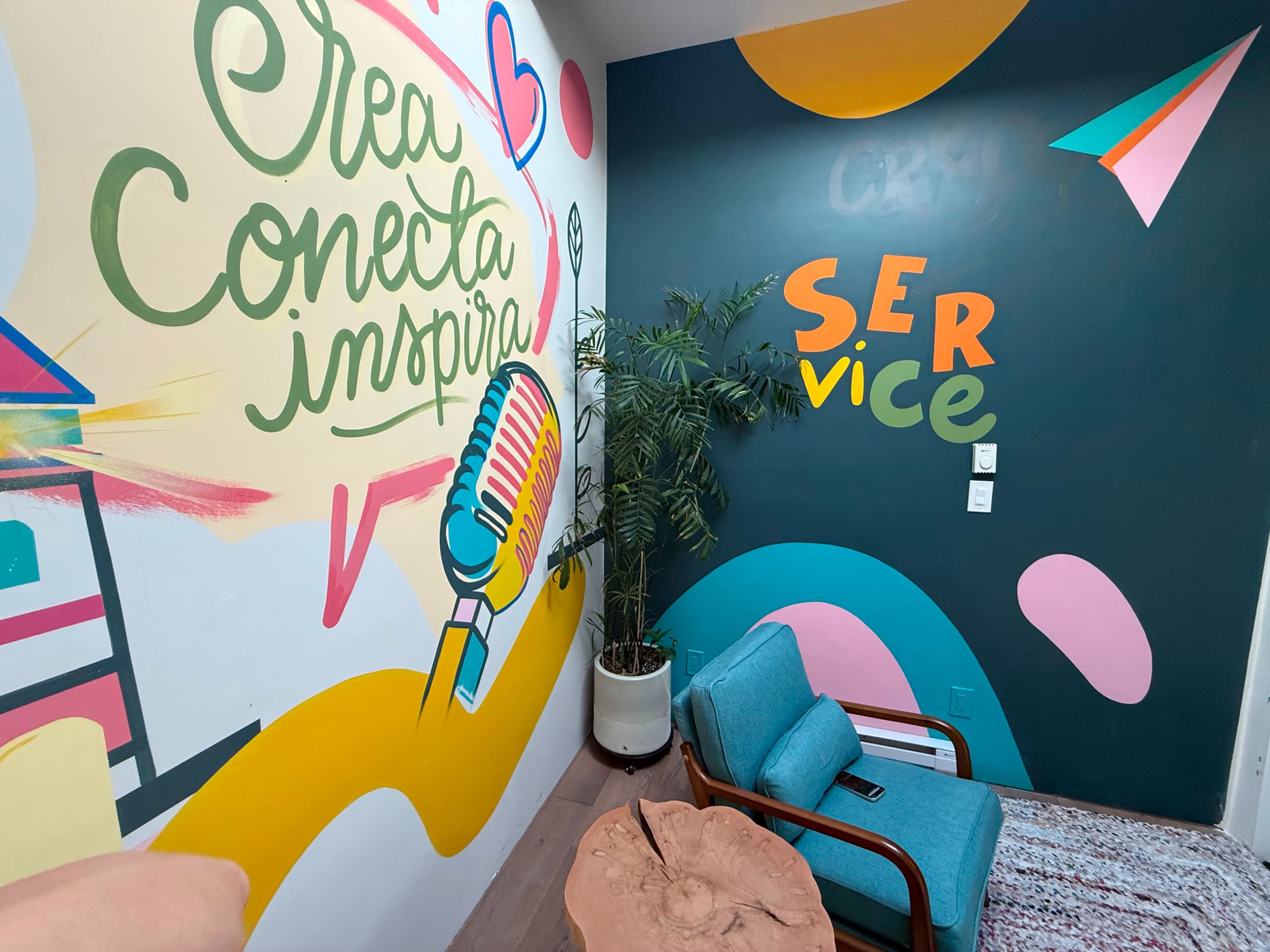 The image shows a colorful room with a mural featuring the words "Crea Conecta Inspira" alongside a microphone, and a corner with a blue chair, wooden table, and a small plant.