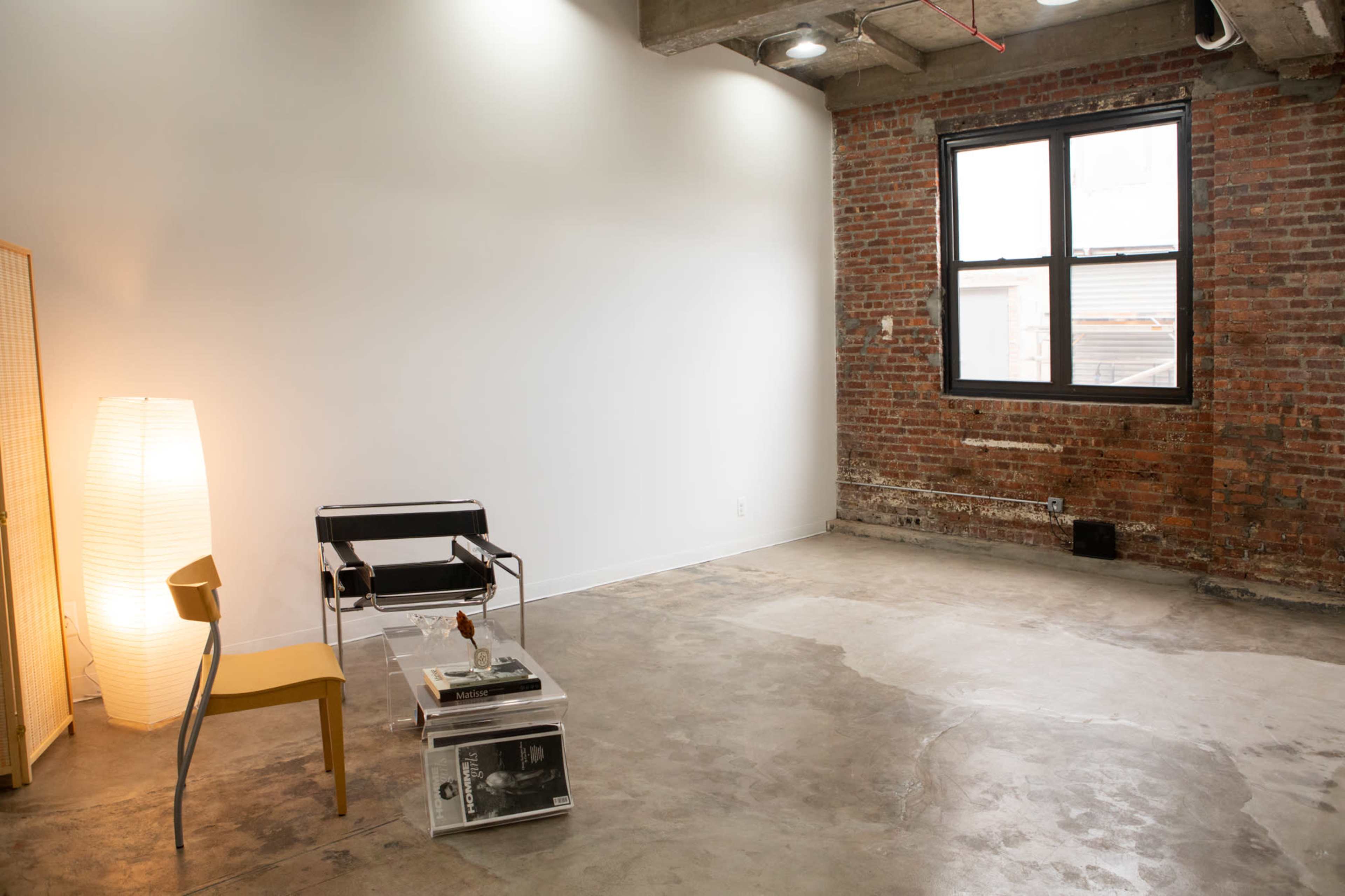 A spacious, industrial-style room with exposed brick walls, a concrete floor, and minimal furniture including a black chair, a yellow chair, and a tall light fixture.