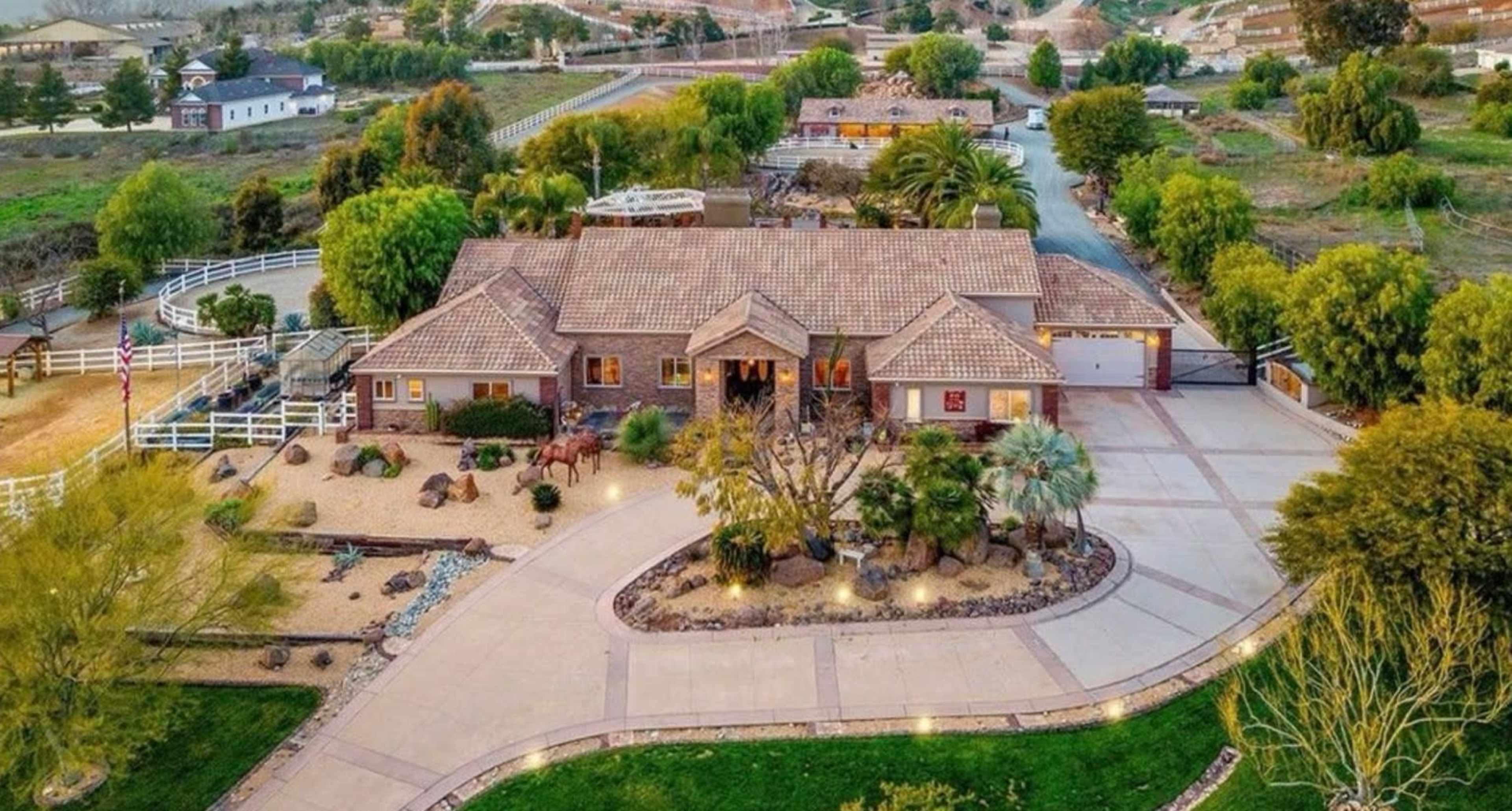 The image shows a large single-story house with a tiled roof, surrounded by a landscaped yard featuring trees, rocks, and a circular driveway.
