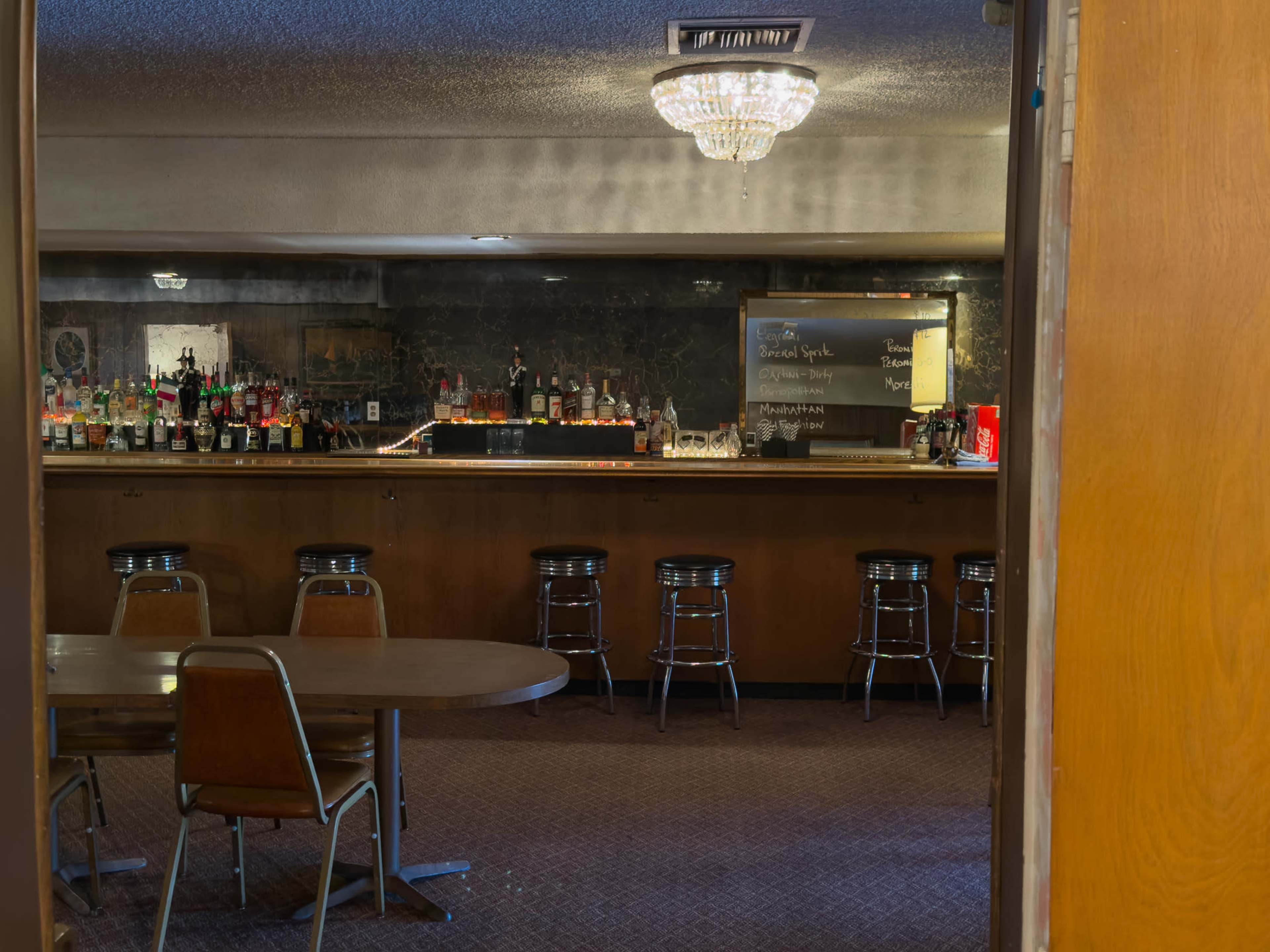 The image shows a dimly lit bar area with a long counter, rows of bottles, and a chandelier overhead, viewed through an open doorway.