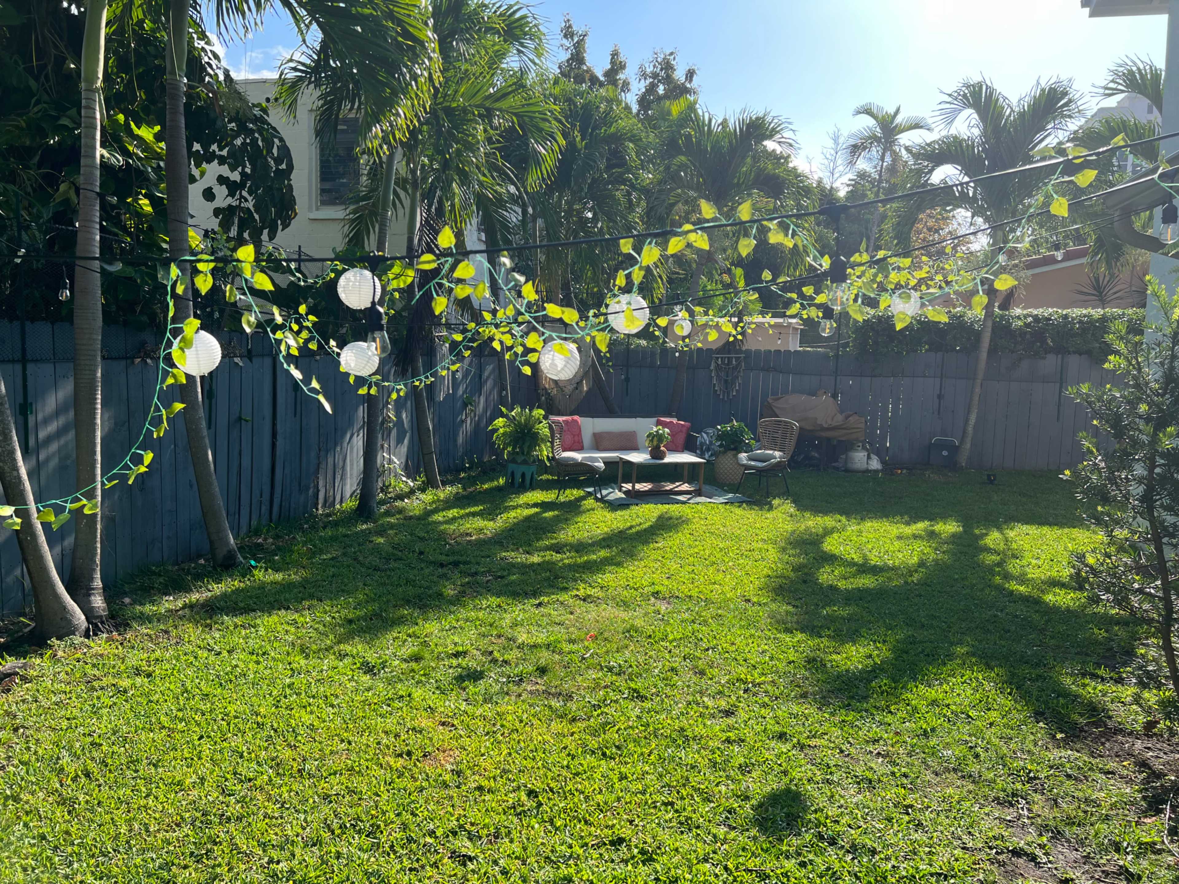 A backyard features a small seating area with pillows, string lights, and greenery, surrounded by palm trees and a wooden fence.