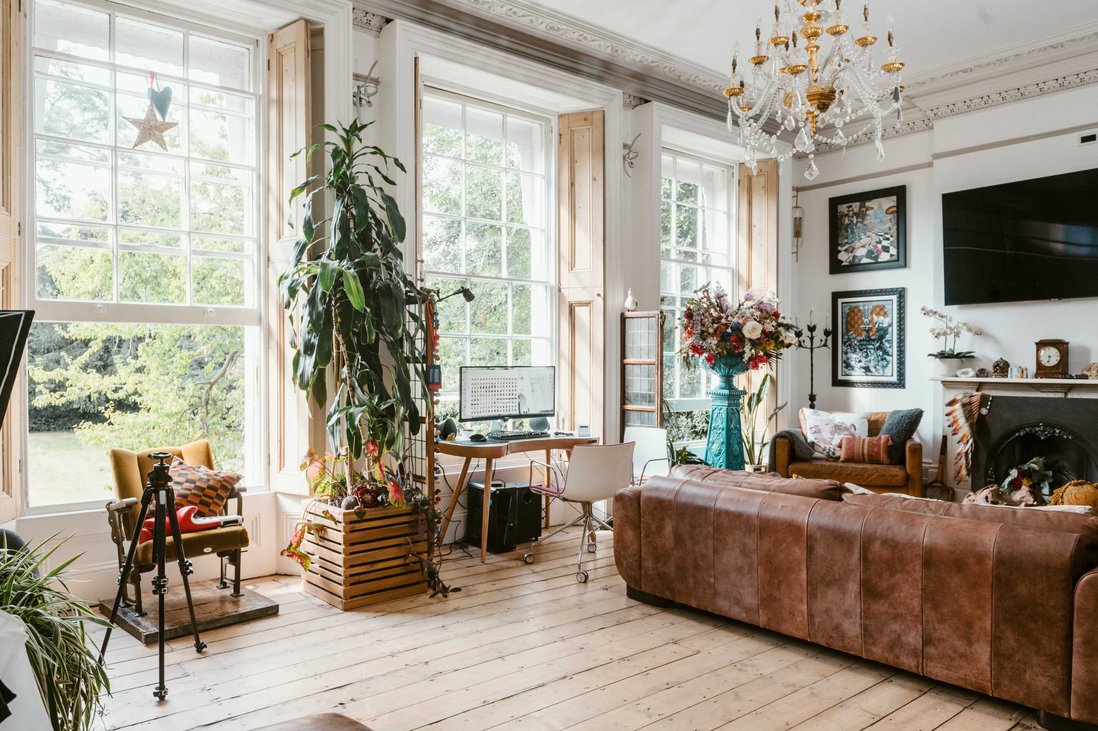 A spacious living room features large windows, a chandelier, a wooden floor, and a mix of modern and vintage furniture, including a desk and a sofa.