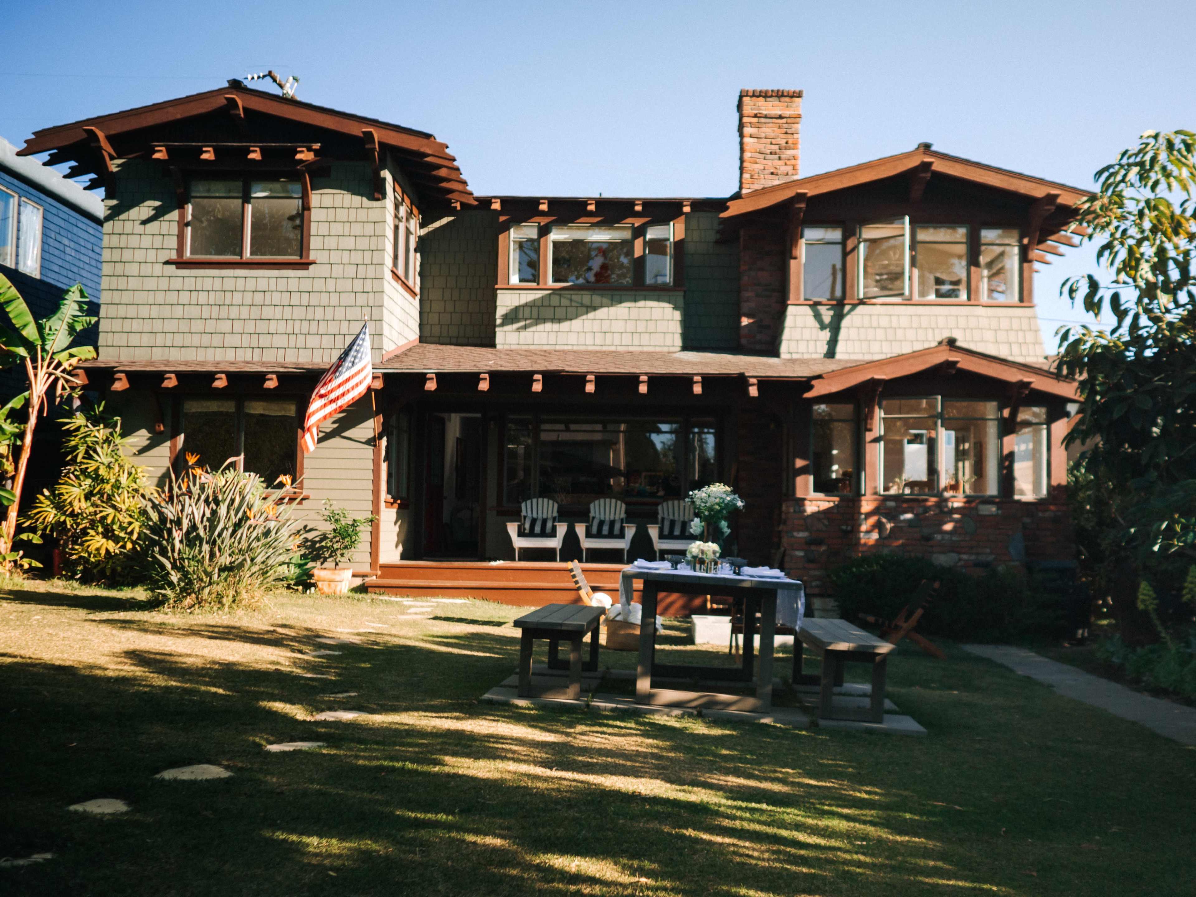 A two-story house with green shingle siding and wooden trim features a front porch with white chairs and a table in the yard, surrounded by shrubs and trees.