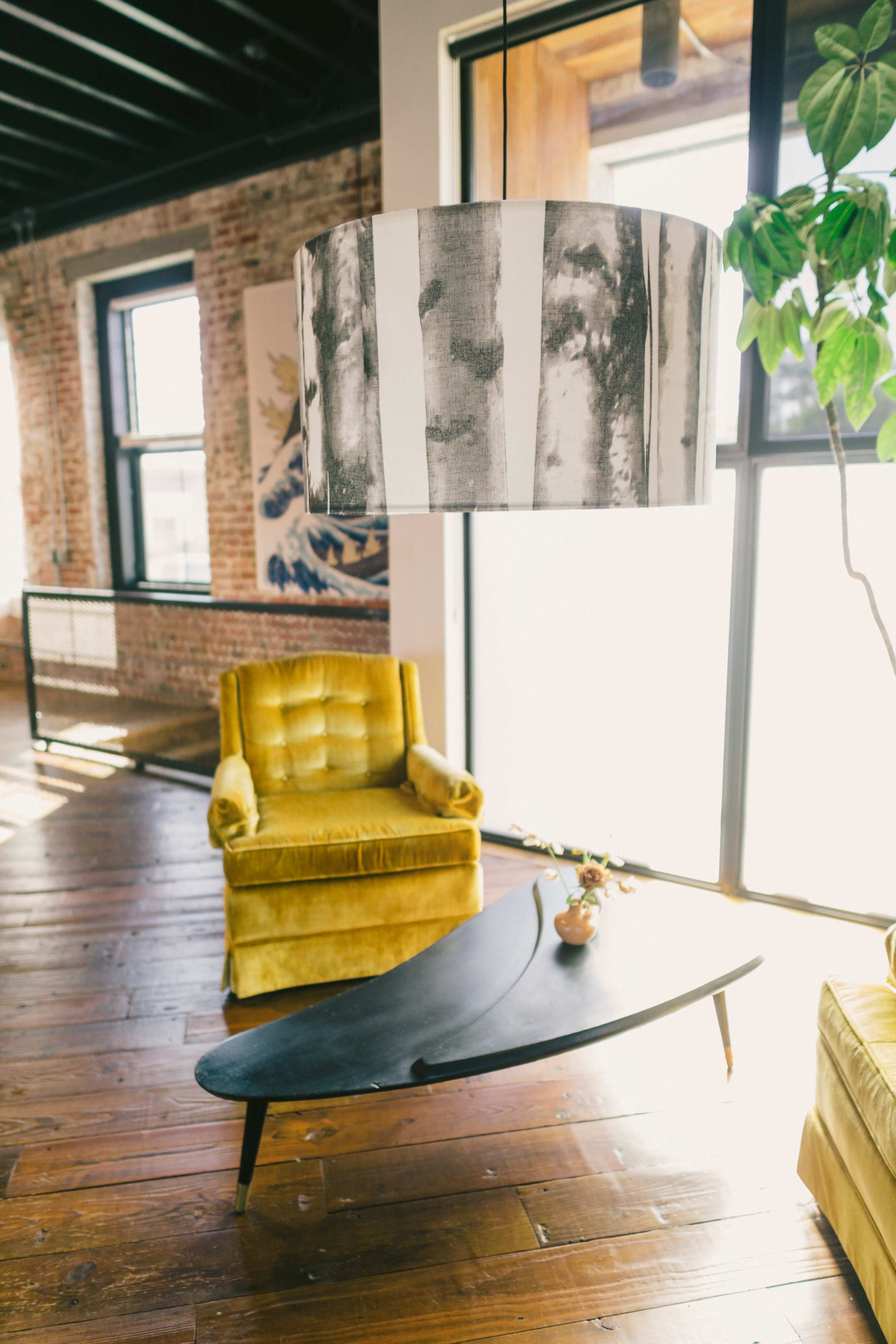 A bright room with a large pendant lamp, a yellow armchair, and a black, curved coffee table, set against a backdrop of exposed brick walls and large windows.
