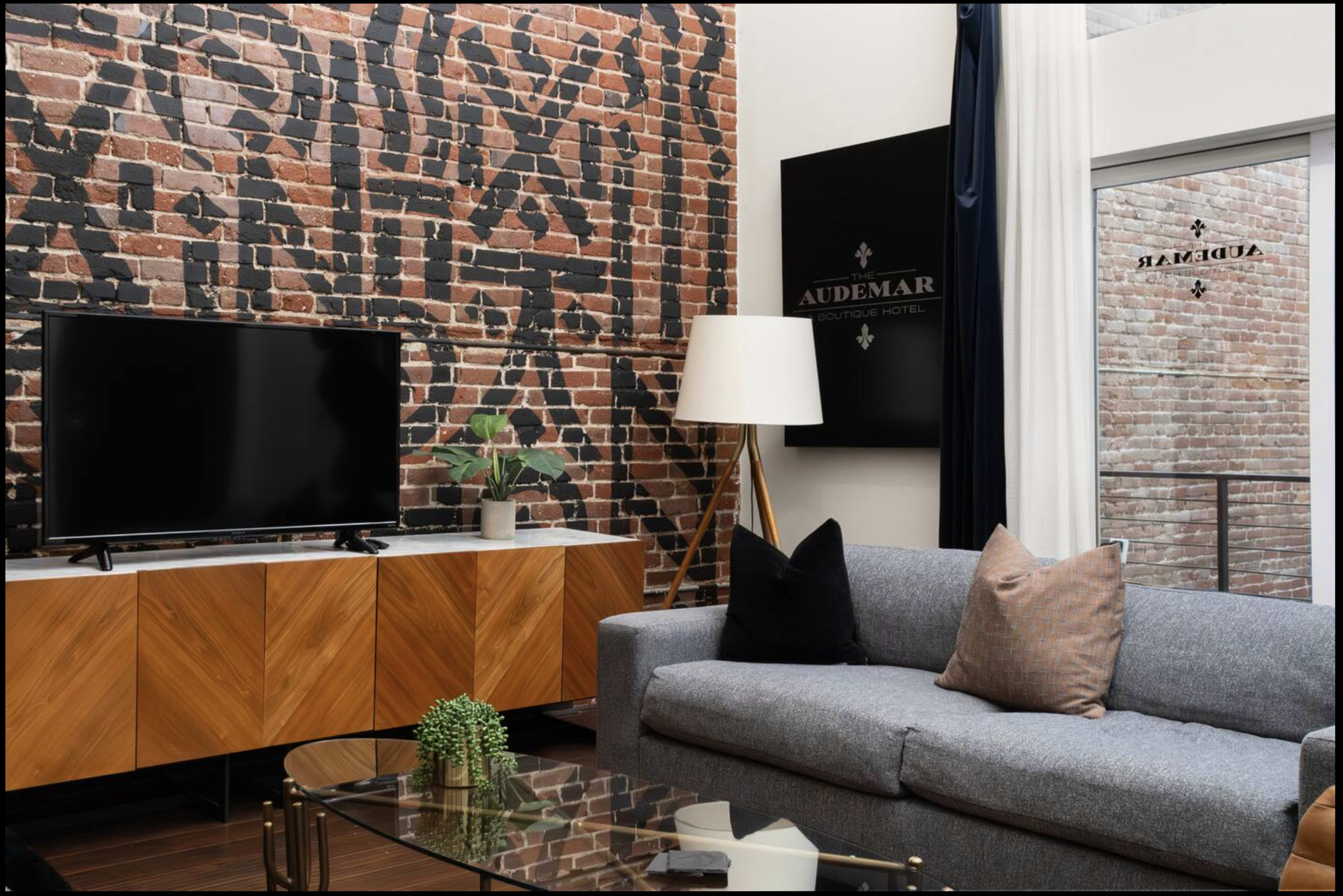 The image shows a modern living space featuring a grey sofa, a glass coffee table, a black television, and a brick accent wall with a large sign.