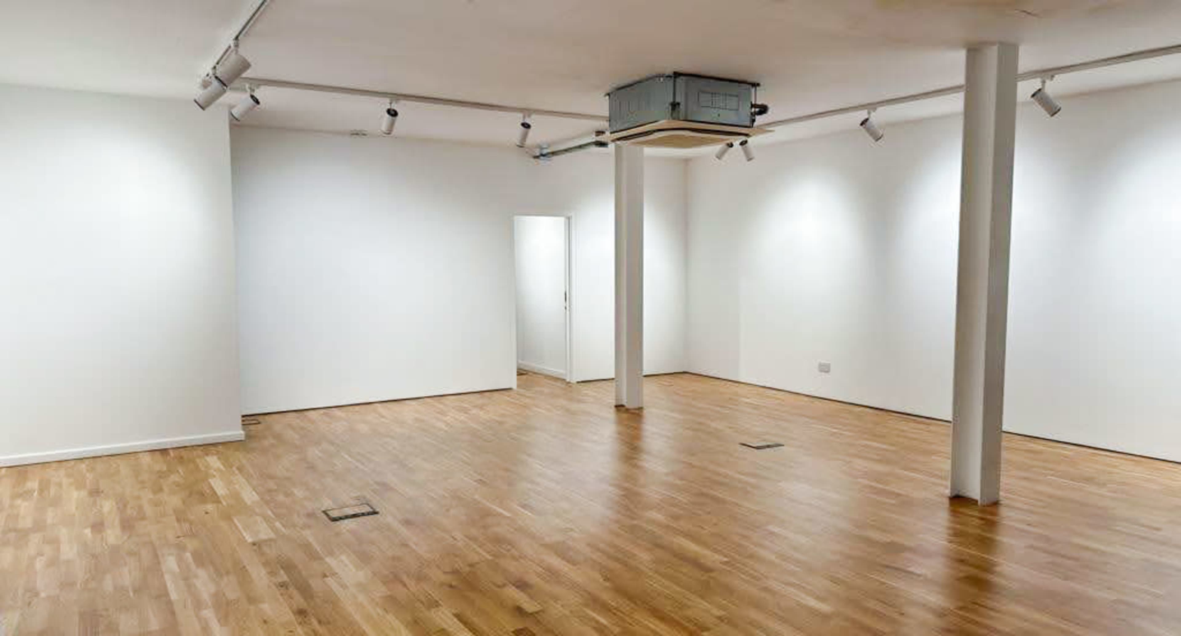 A spacious, empty room with white walls, wooden flooring, and several overhead lights.