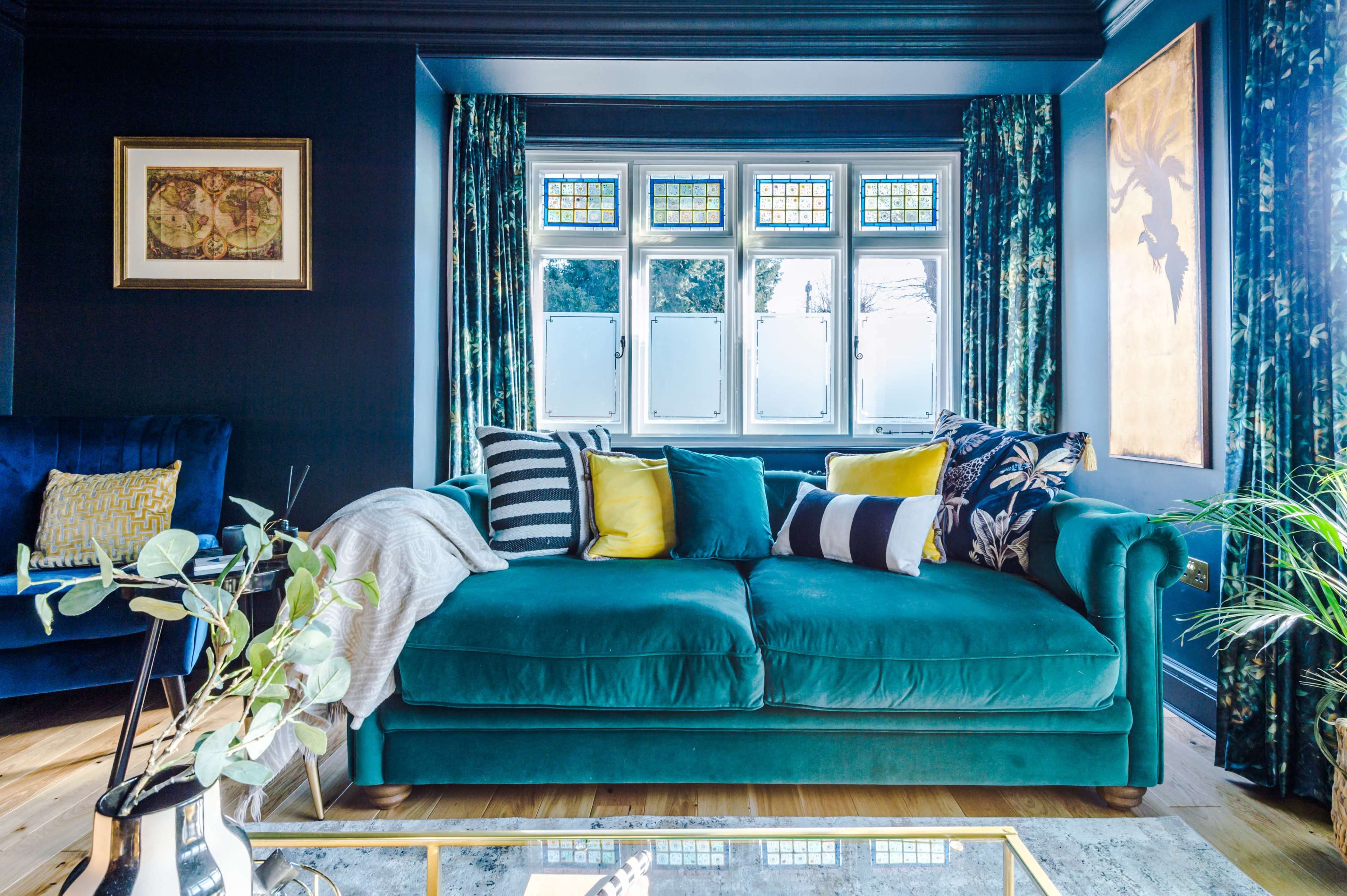 A cozy living room featuring a teal sofa adorned with patterned cushions, a glass coffee table, and large windows with decorative panes.