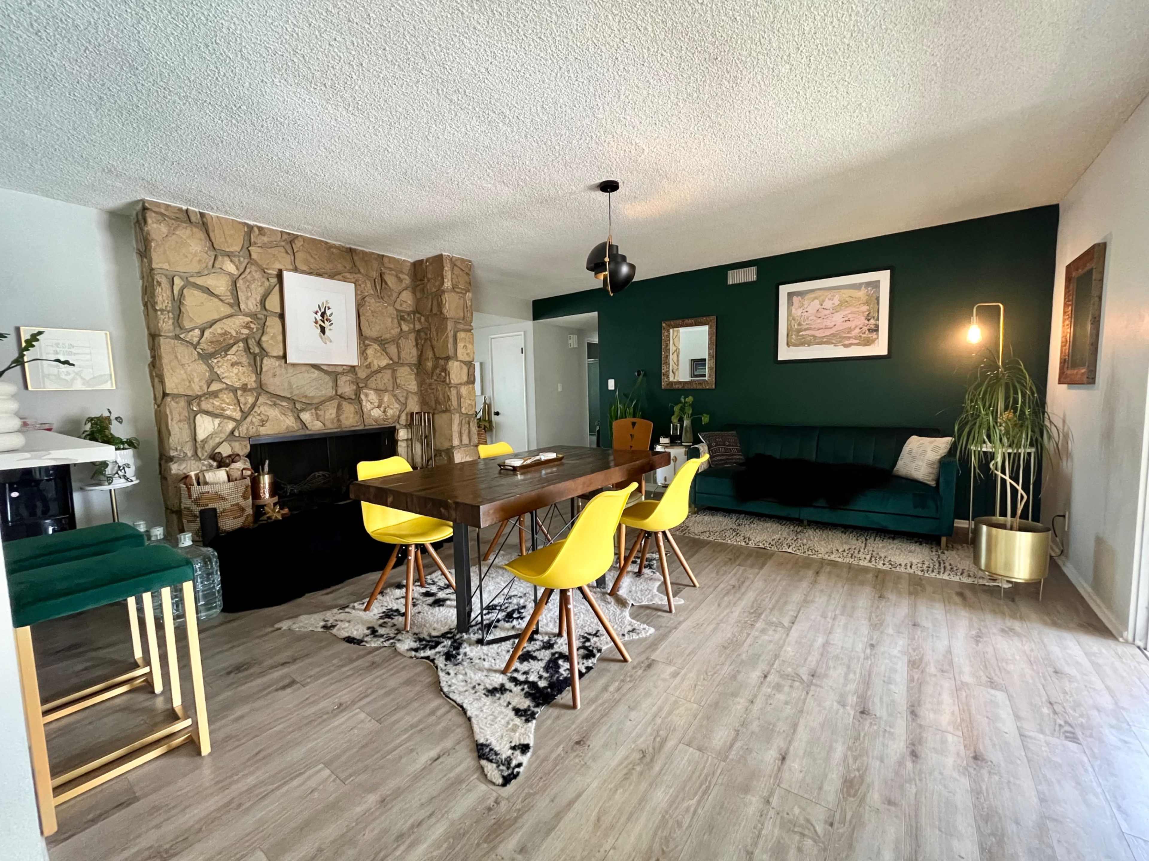 A modern living and dining area with a stone fireplace, a wooden dining table surrounded by yellow chairs, and a dark green accent wall.