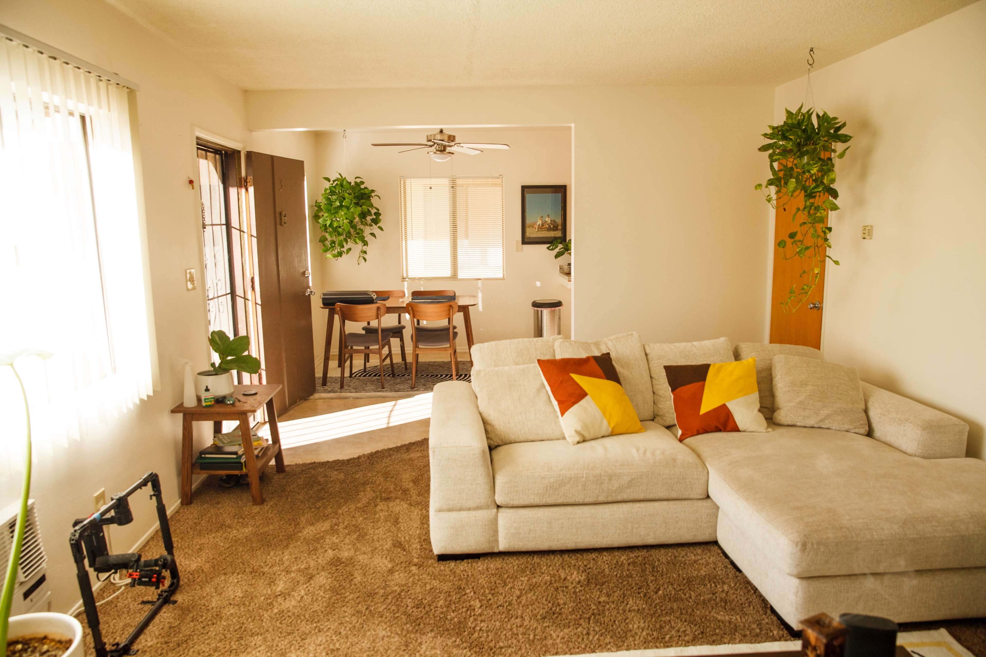 A light-filled living room features a beige sectional sofa with colorful pillows, leading to a dining area with a table and chairs.