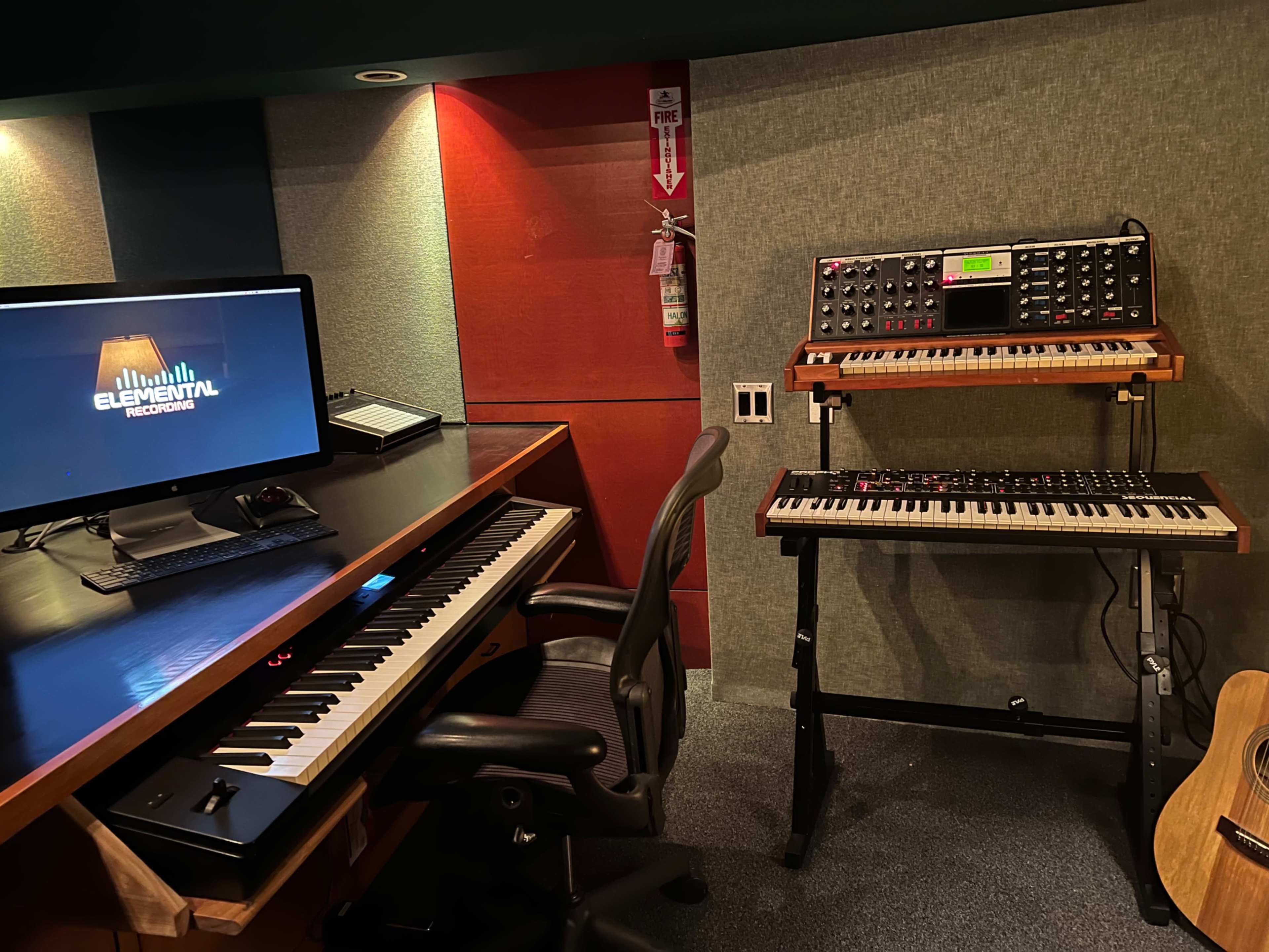 The image shows a music production studio with a computer monitor, a keyboard on a desk, and a synthesizer setup on a stand next to an acoustic guitar.