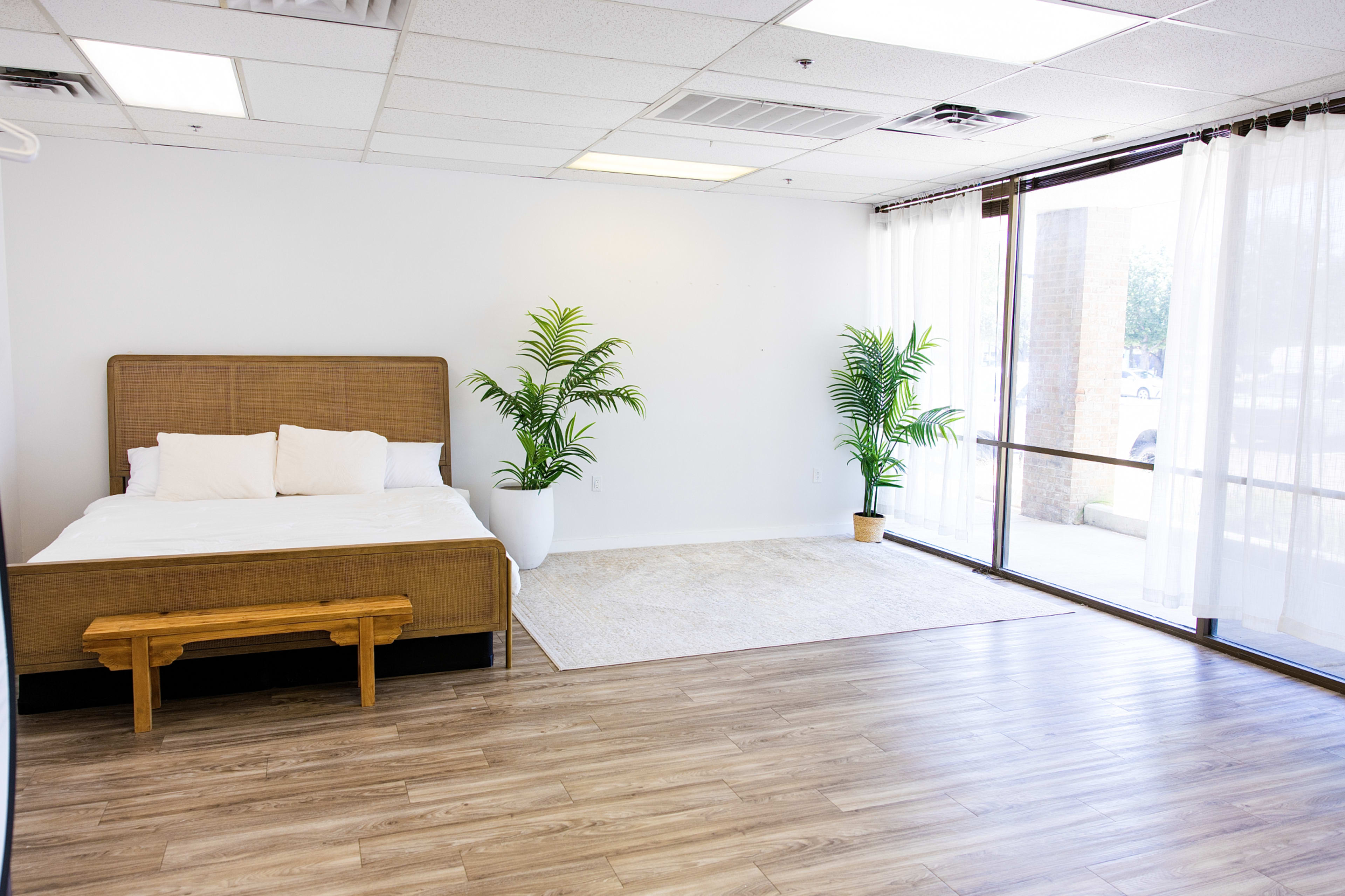 The image shows a minimalist bedroom featuring a wooden bed with white bedding, a small bench at the foot, and two potted plants near a large window with sheer curtains.
