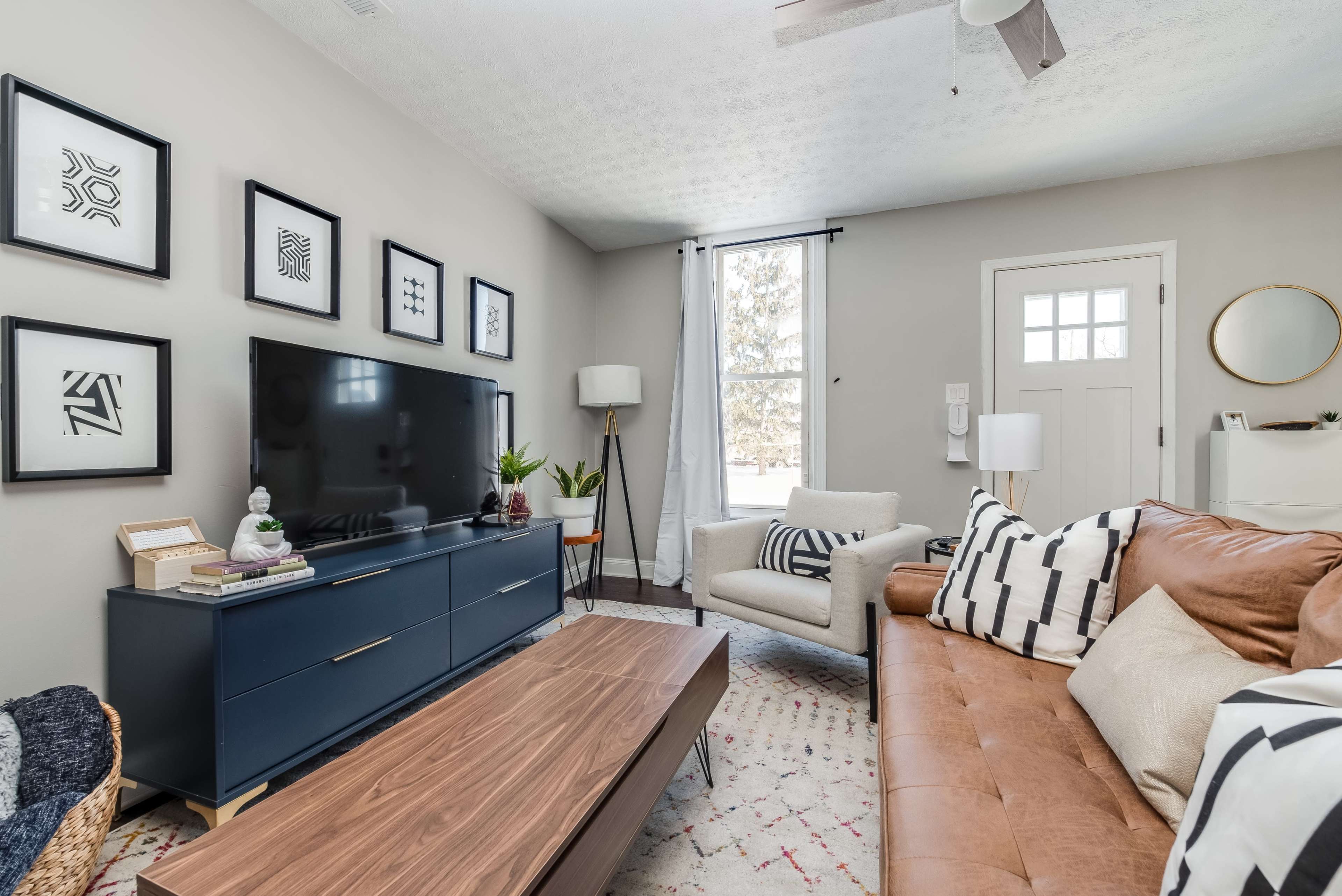 The living room features a combination of a brown leather sofa, a wooden coffee table, and a TV cabinet against a neutral wall, with framed artwork and a tall lamp nearby.