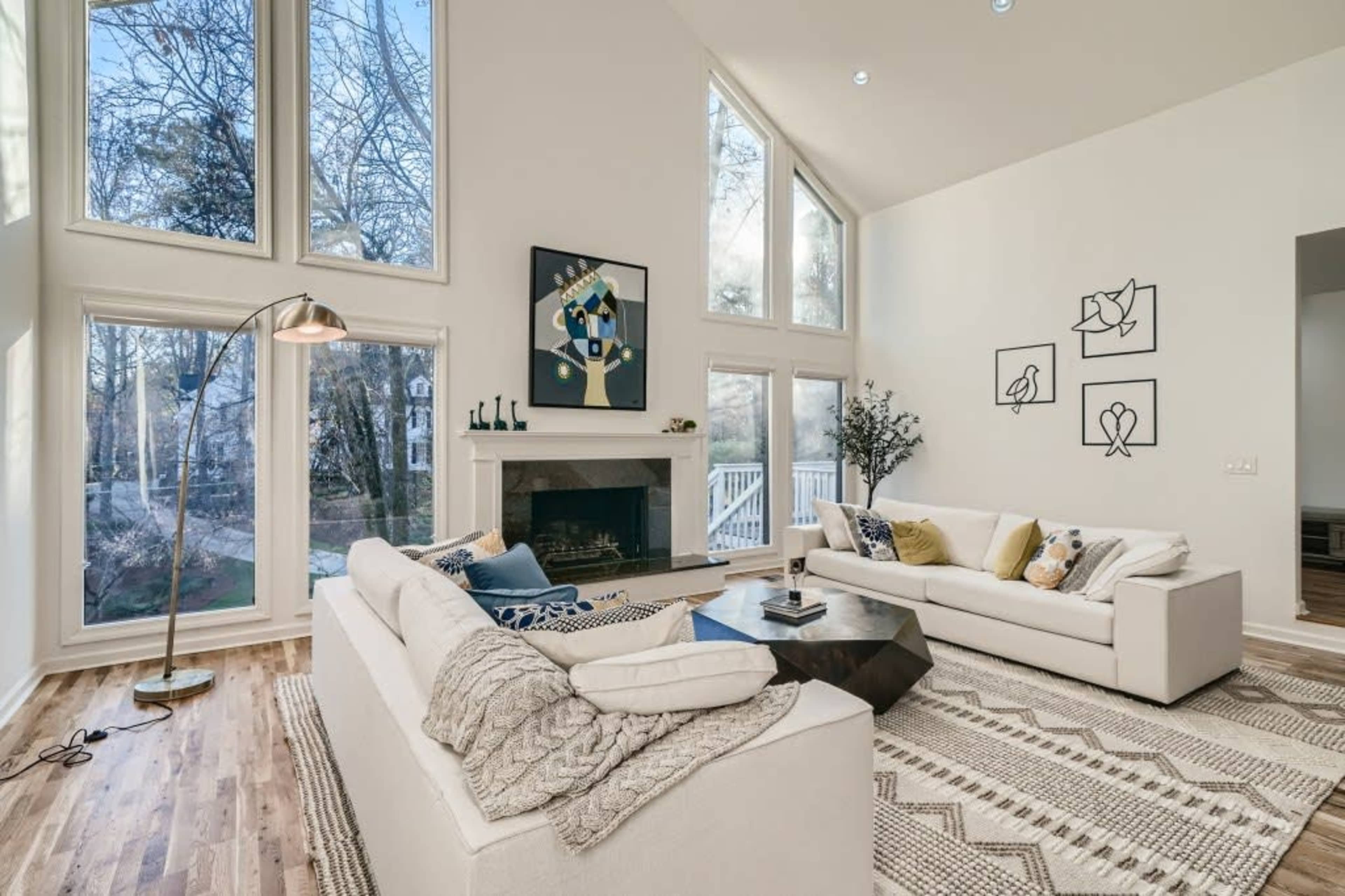 The living room features two white sofas facing a geometric coffee table, with large windows that provide natural light and a view of the outdoors.