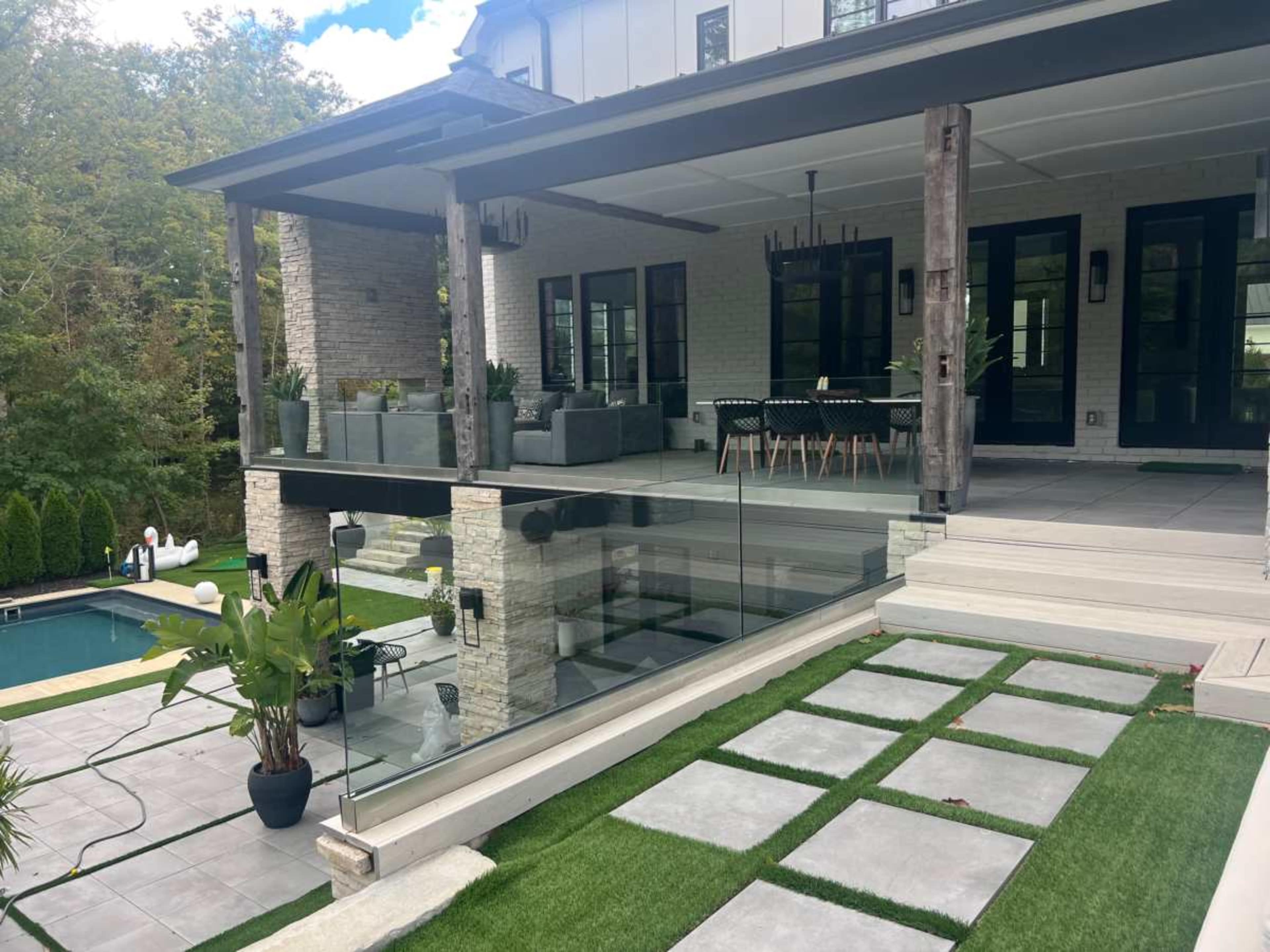The image shows a modern outdoor living space featuring a covered porch with seating, a dining area, and a swimming pool, surrounded by landscaped greenery and stone pathways.