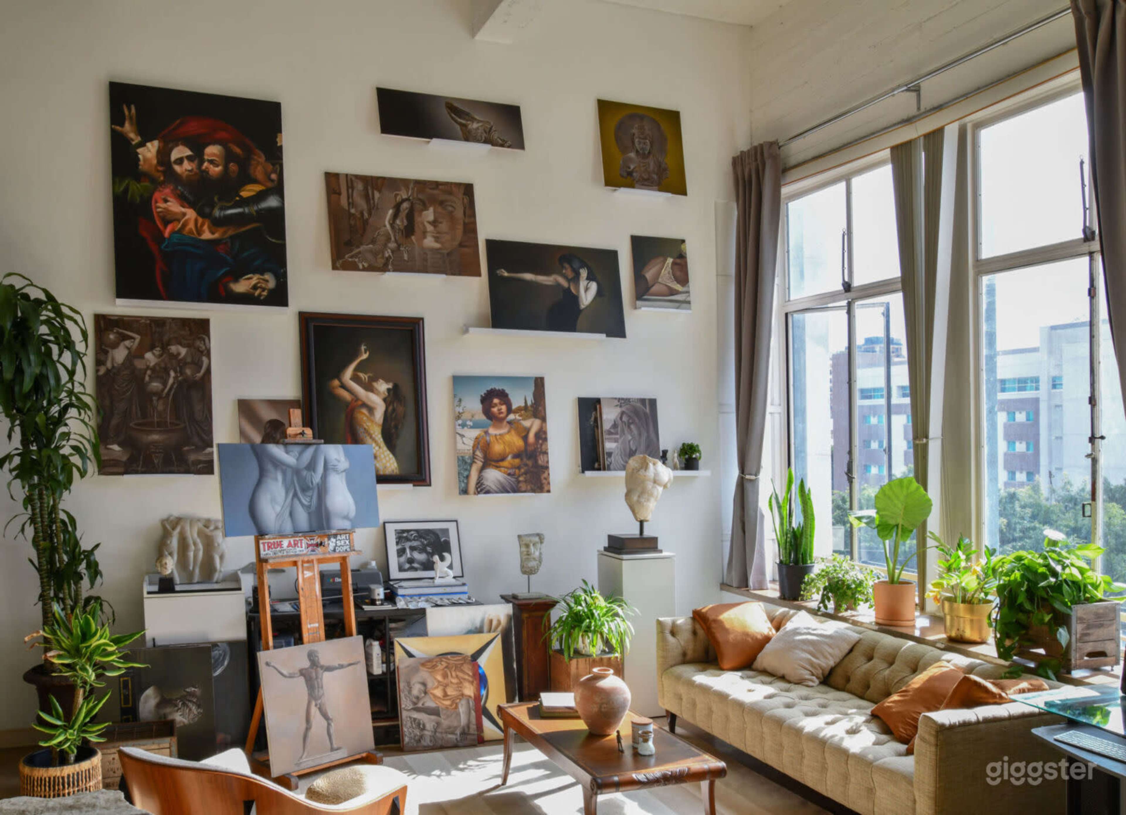 The room features a modern art studio with a gallery wall displaying various paintings and sculptures, along with a comfortable sofa and several indoor plants.