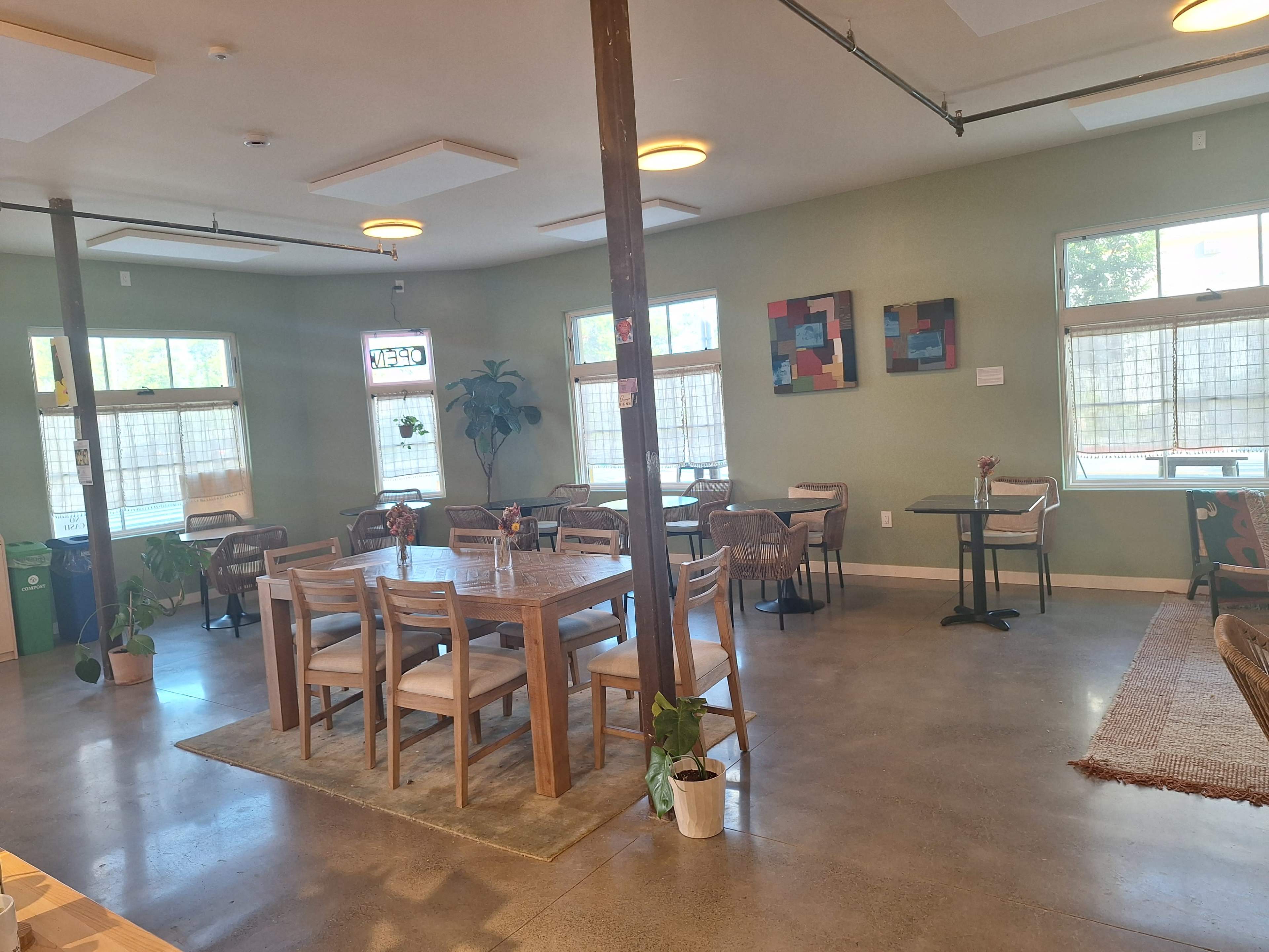 The image shows a spacious dining area with tables and chairs arranged neatly, featuring large windows and artworks on the walls.