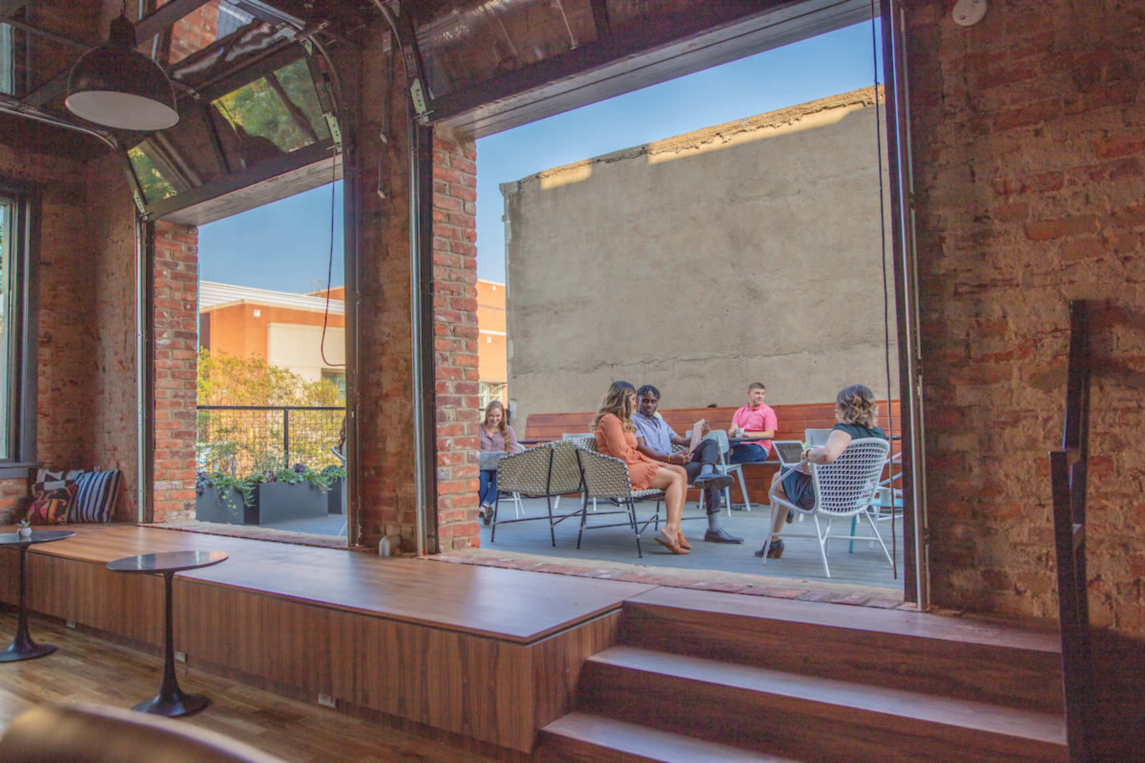 A group of people sits in a casual setting on a patio, framed by large open windows in a brick building.