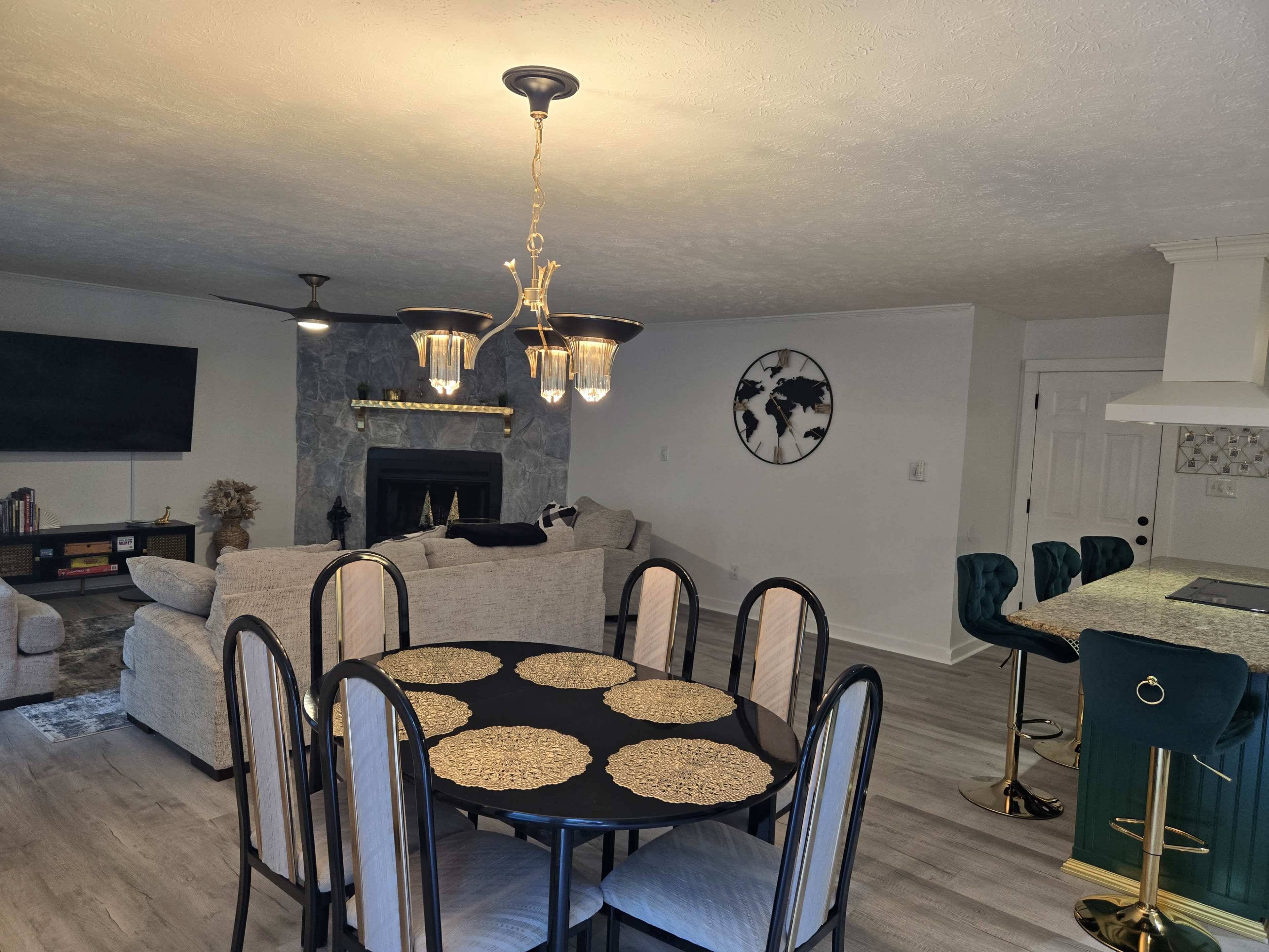 The image shows a modern open-concept living and dining area featuring a round dining table with black chairs, a light fixture above, a gray sofa, a television, and a bar area with green stools.