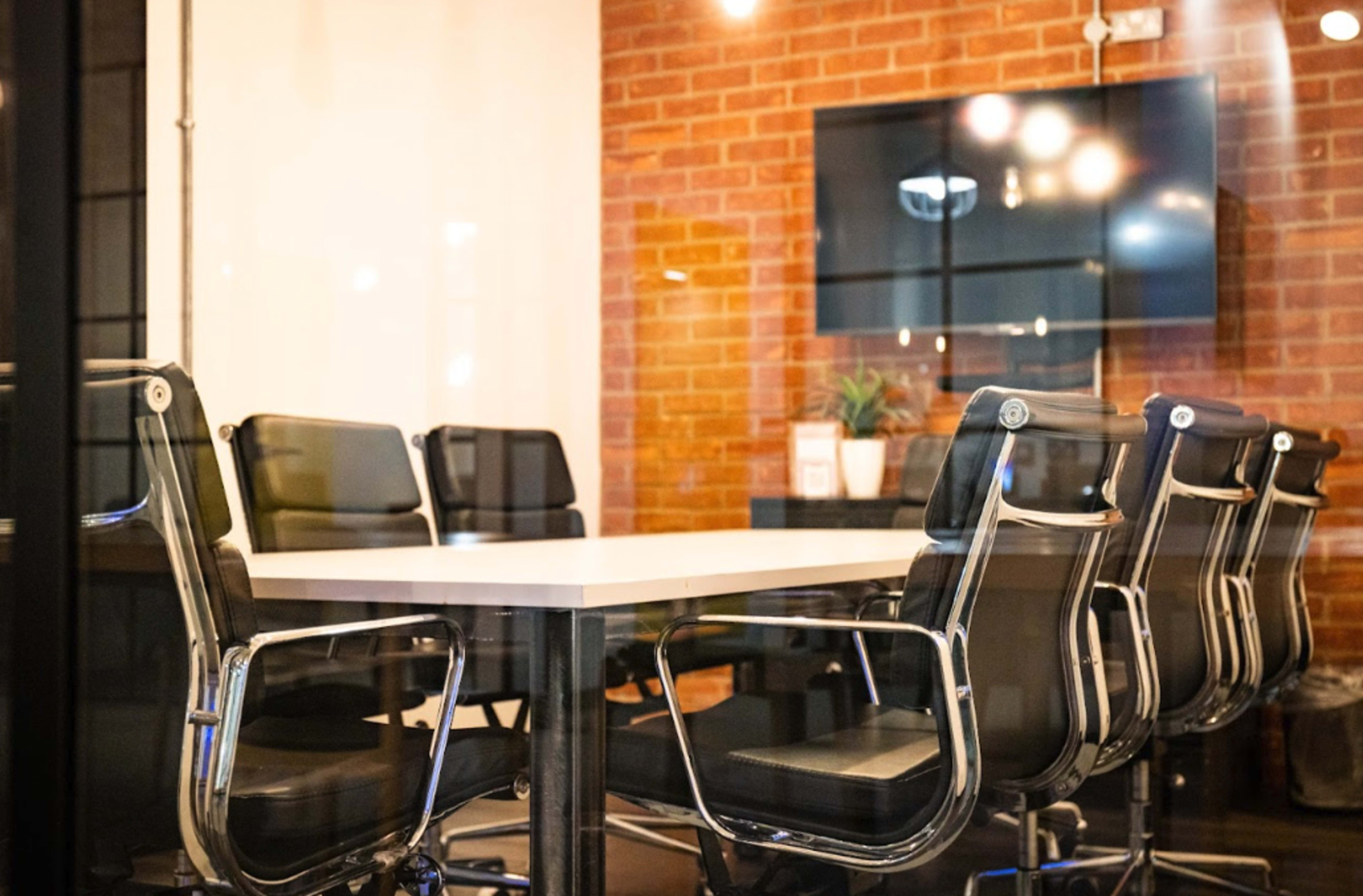 A conference room with a large table surrounded by several black office chairs, featuring a brick wall and a wall-mounted screen.