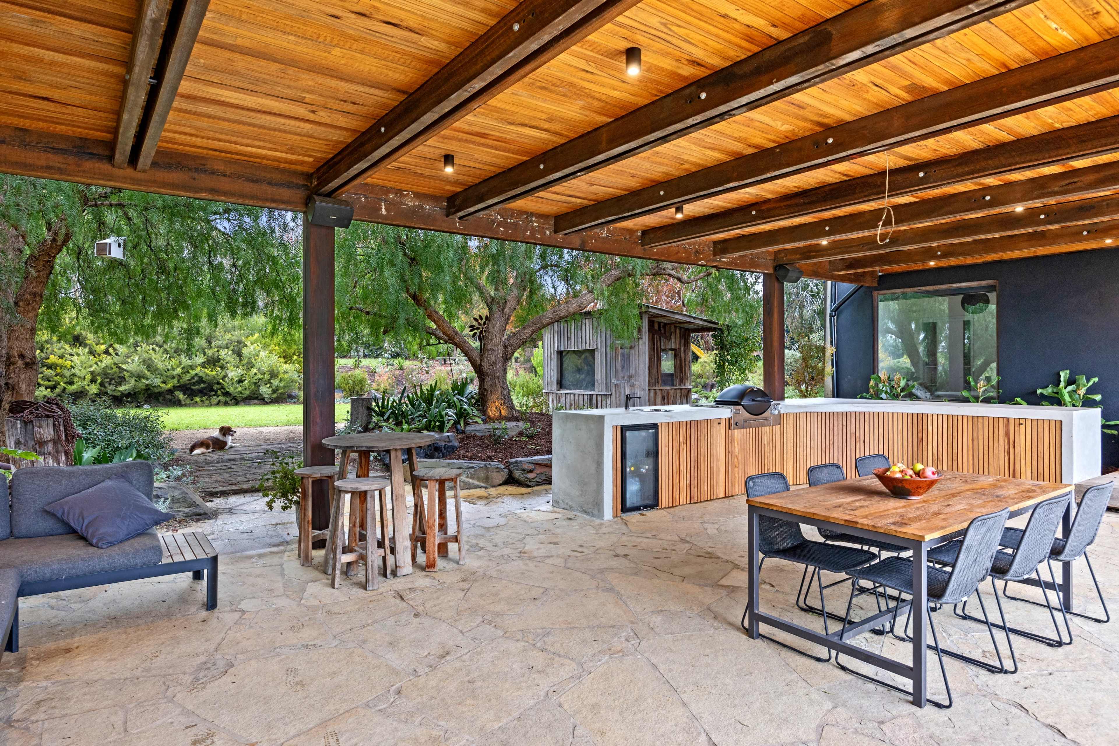 A covered outdoor patio features a wooden dining table and chairs, a kitchen area, and a stone floor, surrounded by greenery and trees.