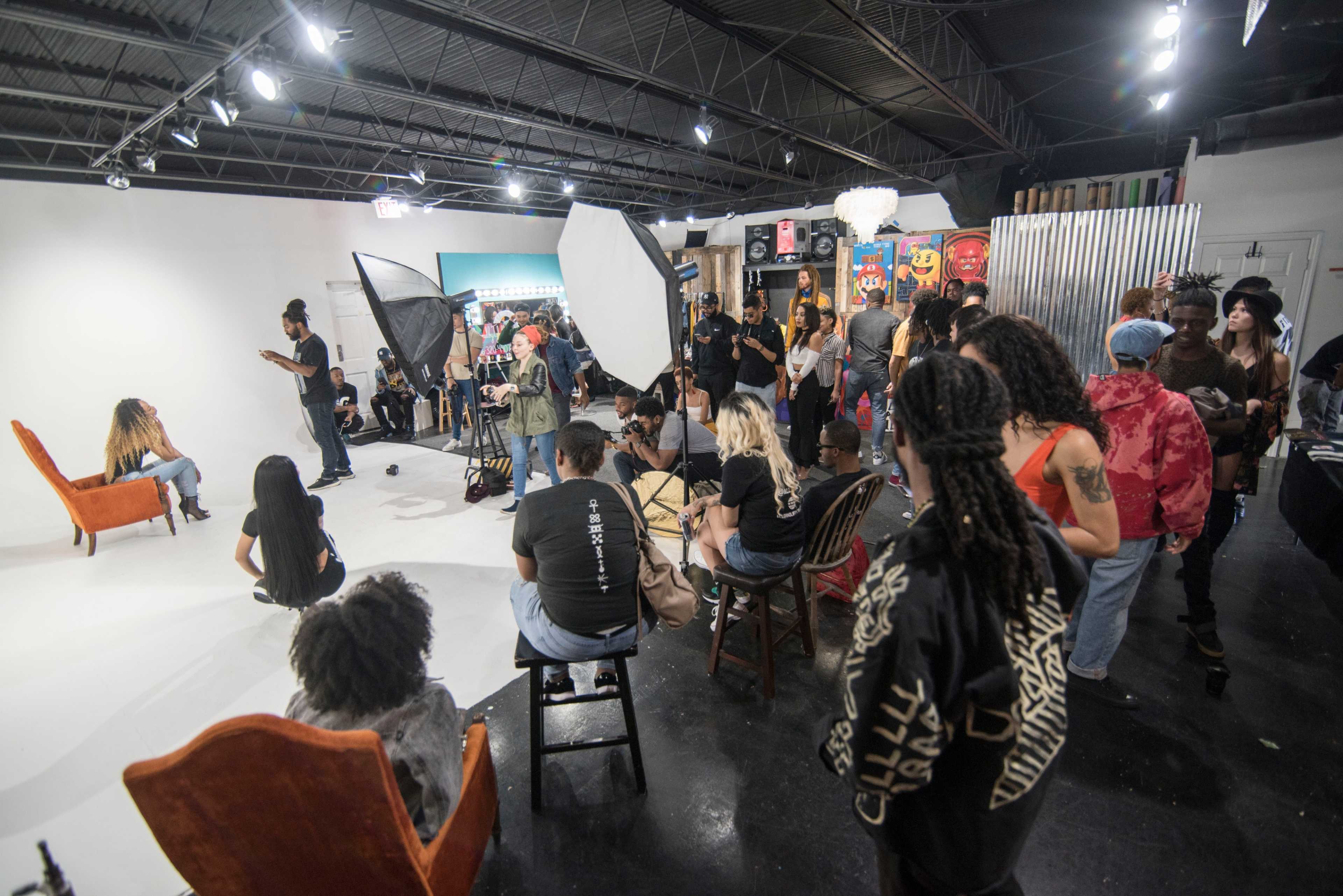 A group of people observes a photo shoot taking place in a studio with a white backdrop and various props.