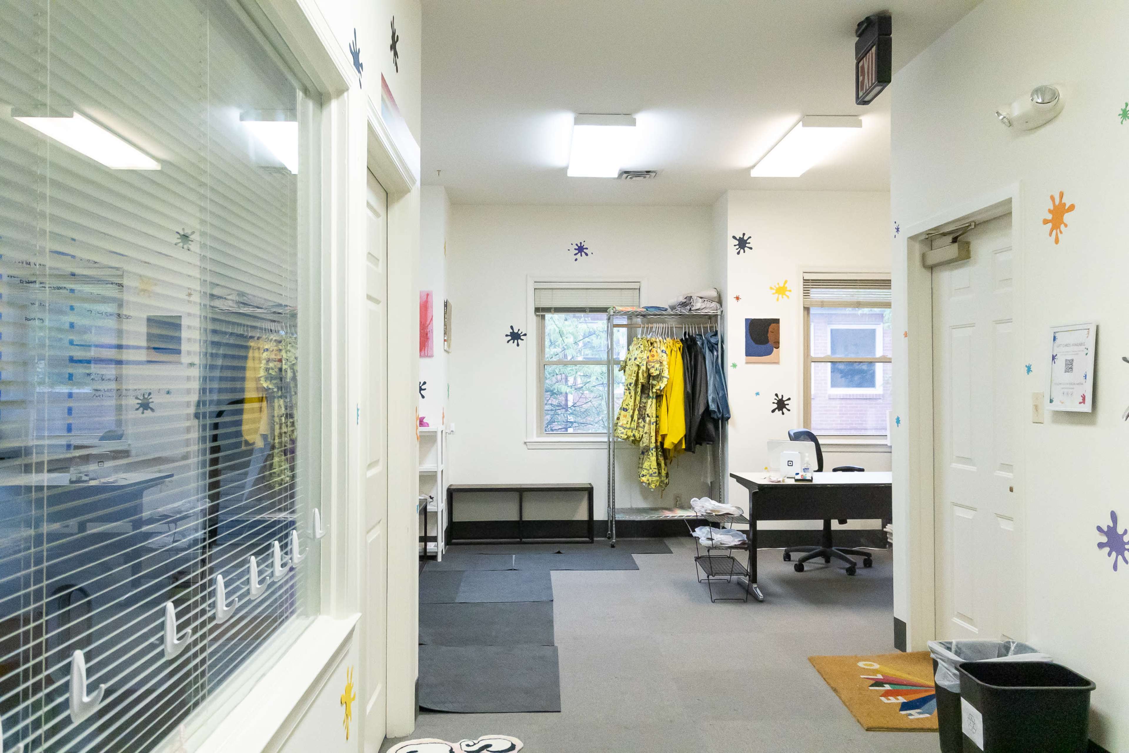 A brightly lit hallway with two doors, a view into a room with clothing on display, and an office space at the end.