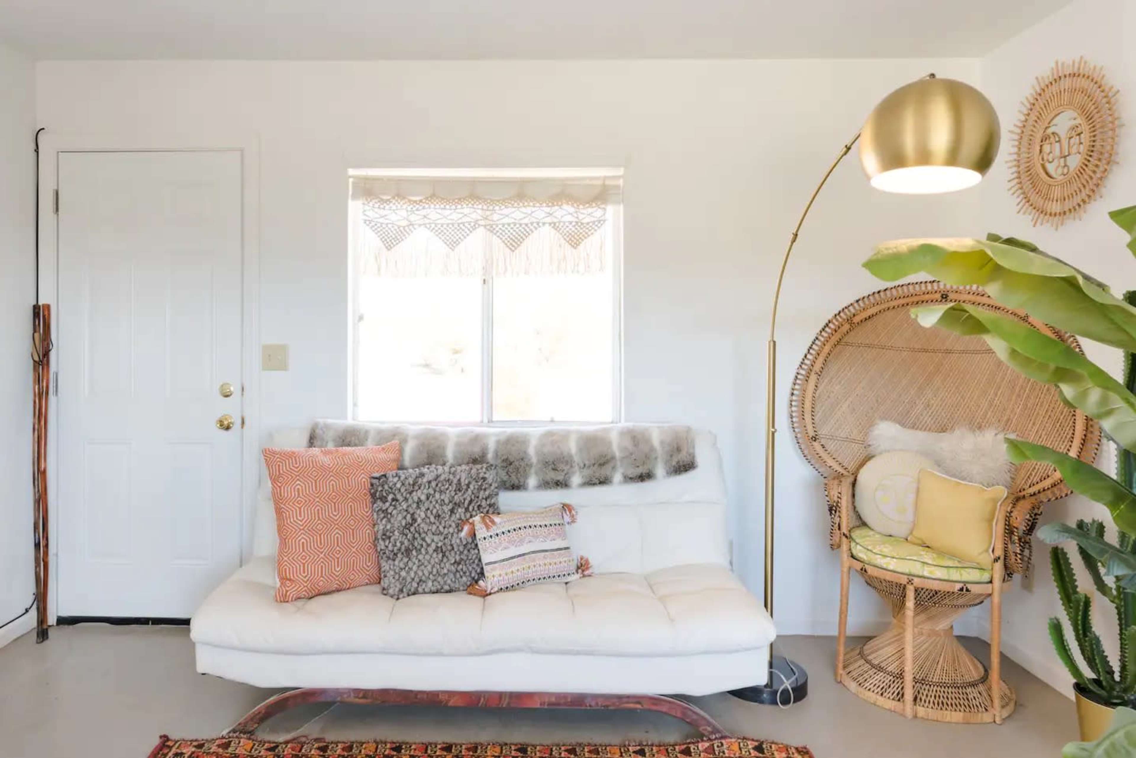 A bright room featuring a white sofa with decorative pillows, a large rattan chair, a standing lamp, and a potted plant, all arranged near a window.