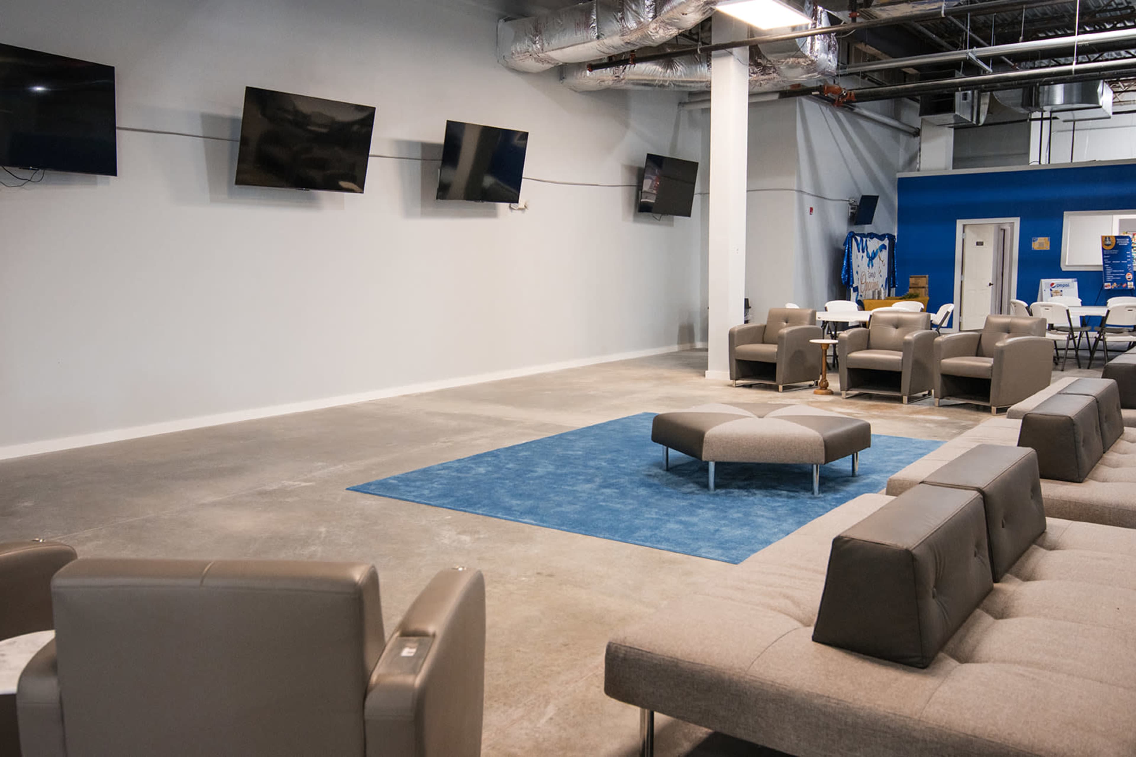 The image shows a spacious lounge area with several wall-mounted televisions, grey seating arrangements, and a large blue rug on a concrete floor.