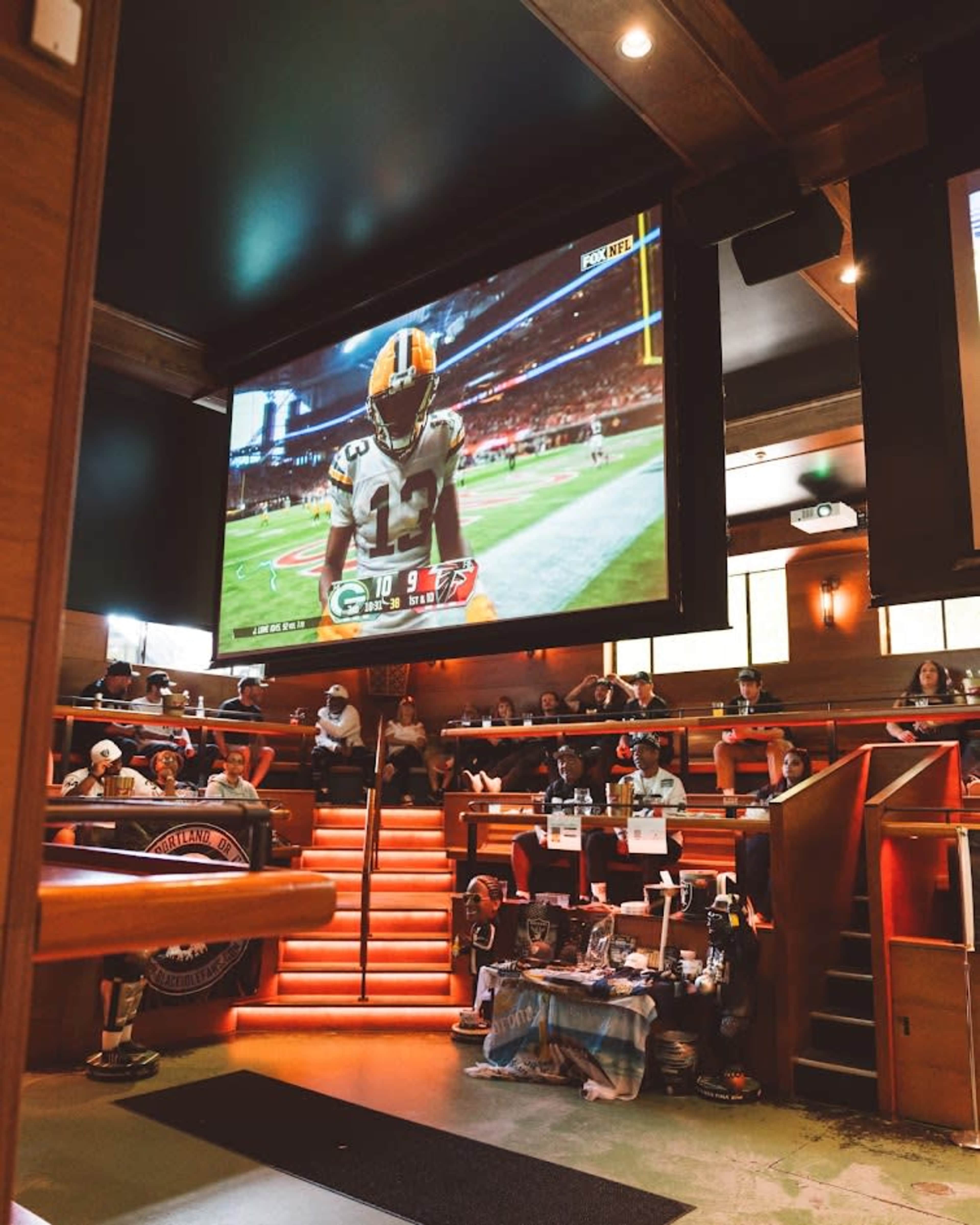 The image shows a sports bar with a large screen displaying a football game, where a player from the Green Bay Packers is highlighted, while patrons watch and sit in an elevated seating area.