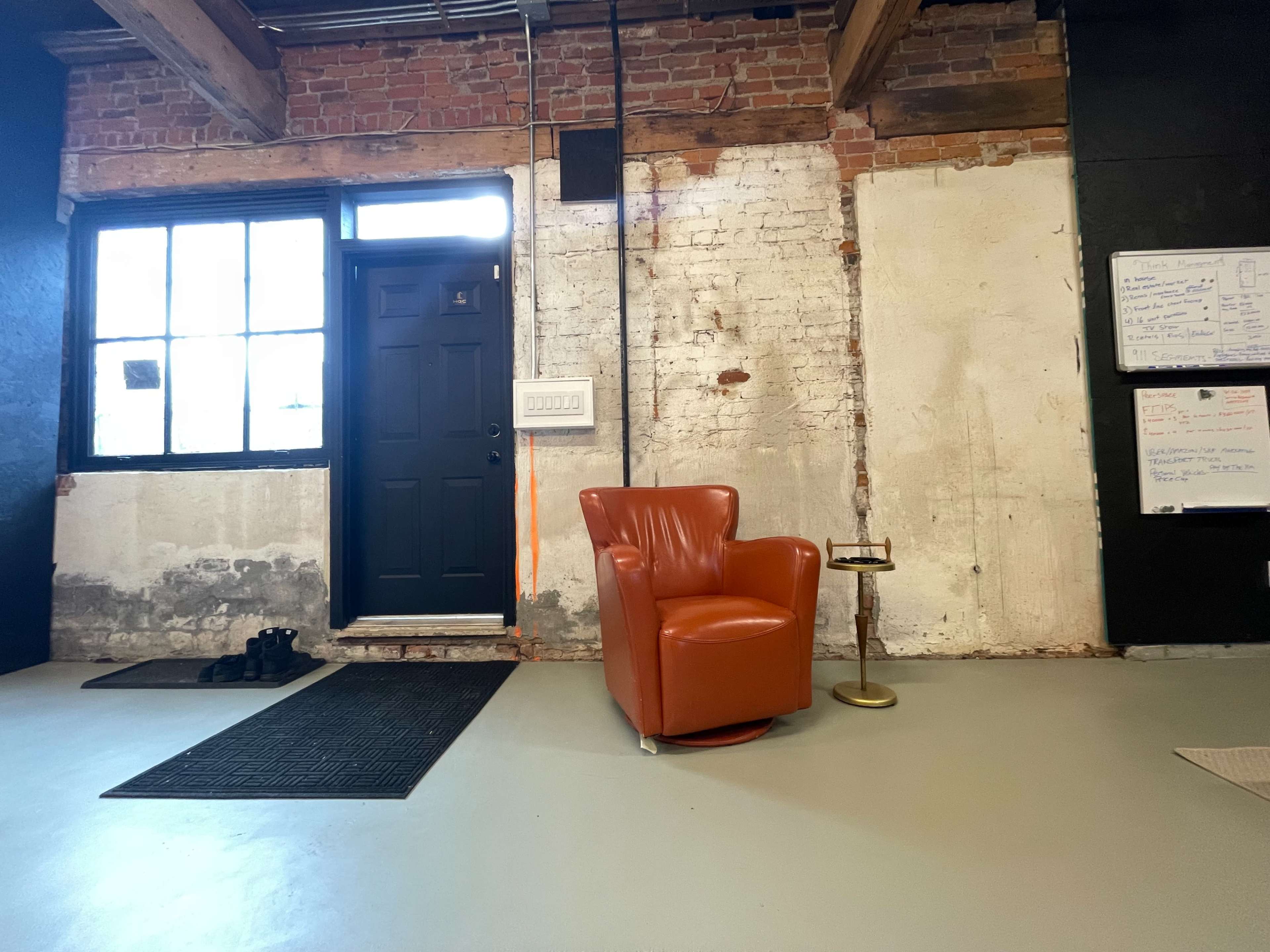 A minimalist interior space featuring a dark door, a vintage orange armchair, a small side table, and exposed brick walls.
